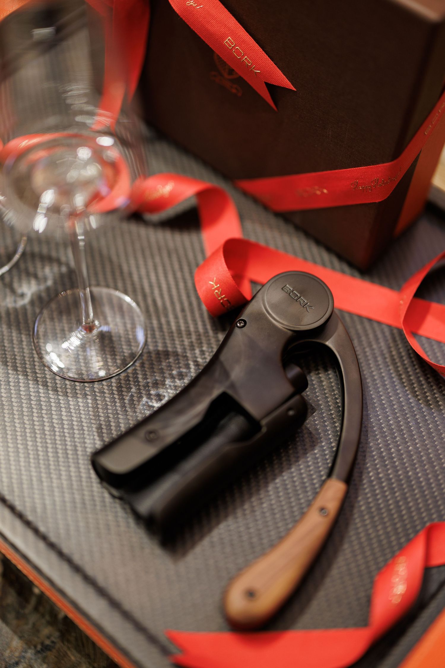 Close-up of wine accessories including a corkscrew, red ribbon, and wine glass on a textured surface.