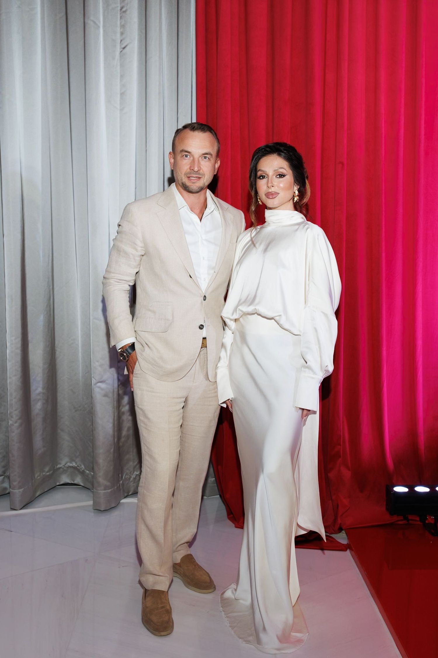 Two people pose in matching white outfits against a backdrop with red and gray curtains.