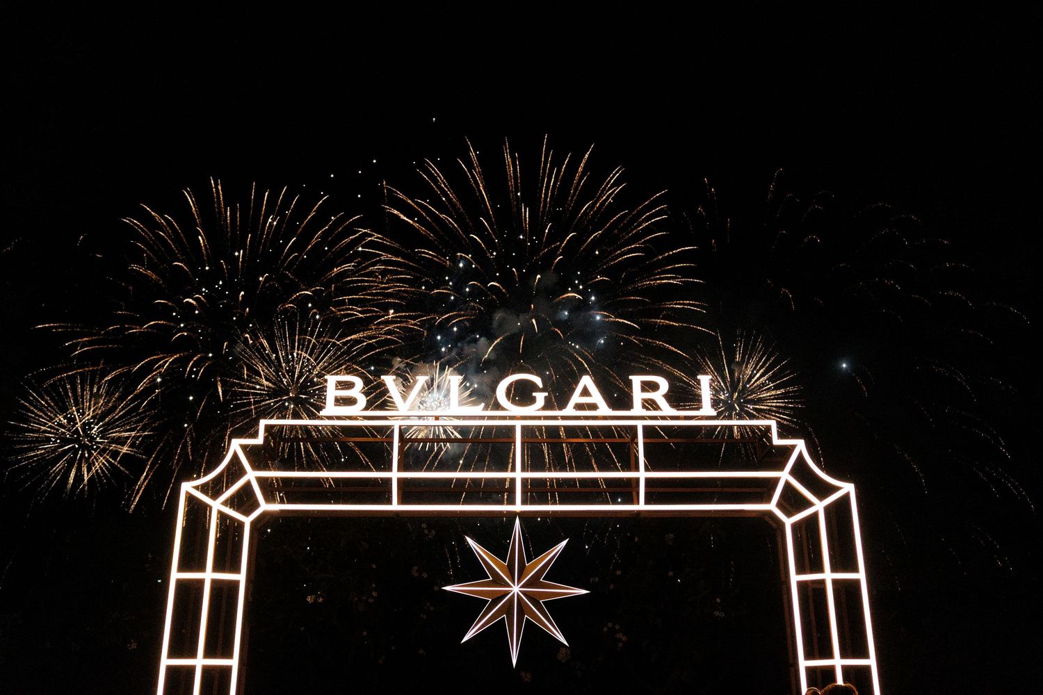 Fireworks explode in the night sky above illuminated BVLGARI signage on a building facade.