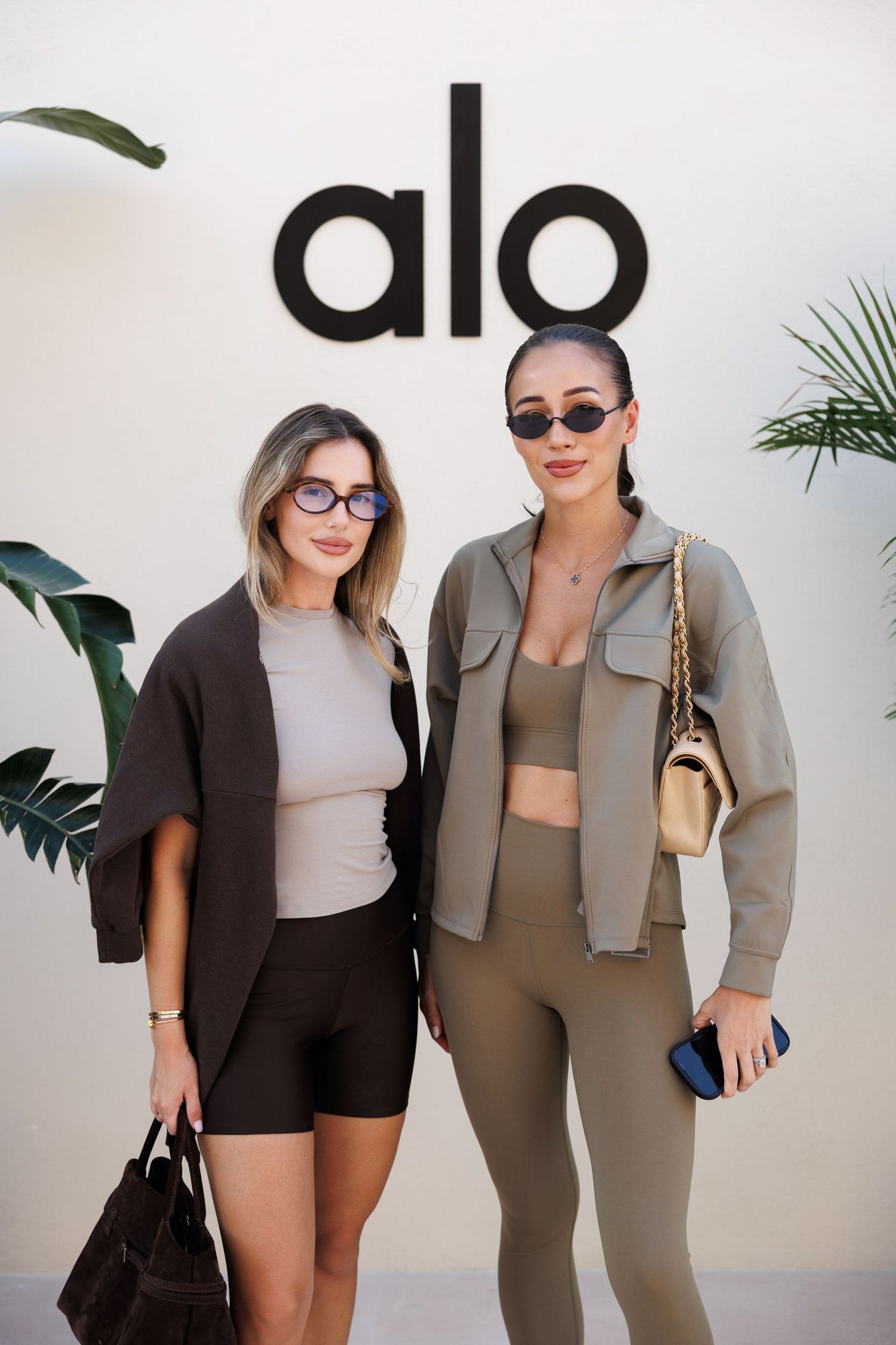 Two women modeling athleisure outfits in front of a white wall with 'alo' signage.