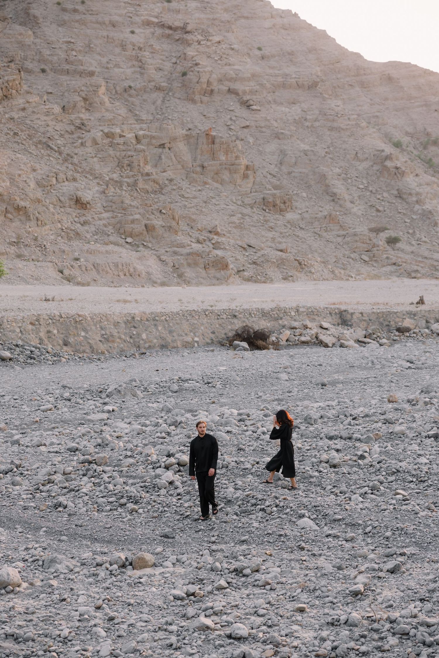 Two people walking through a desert landscape with rocky cliffs in the background.