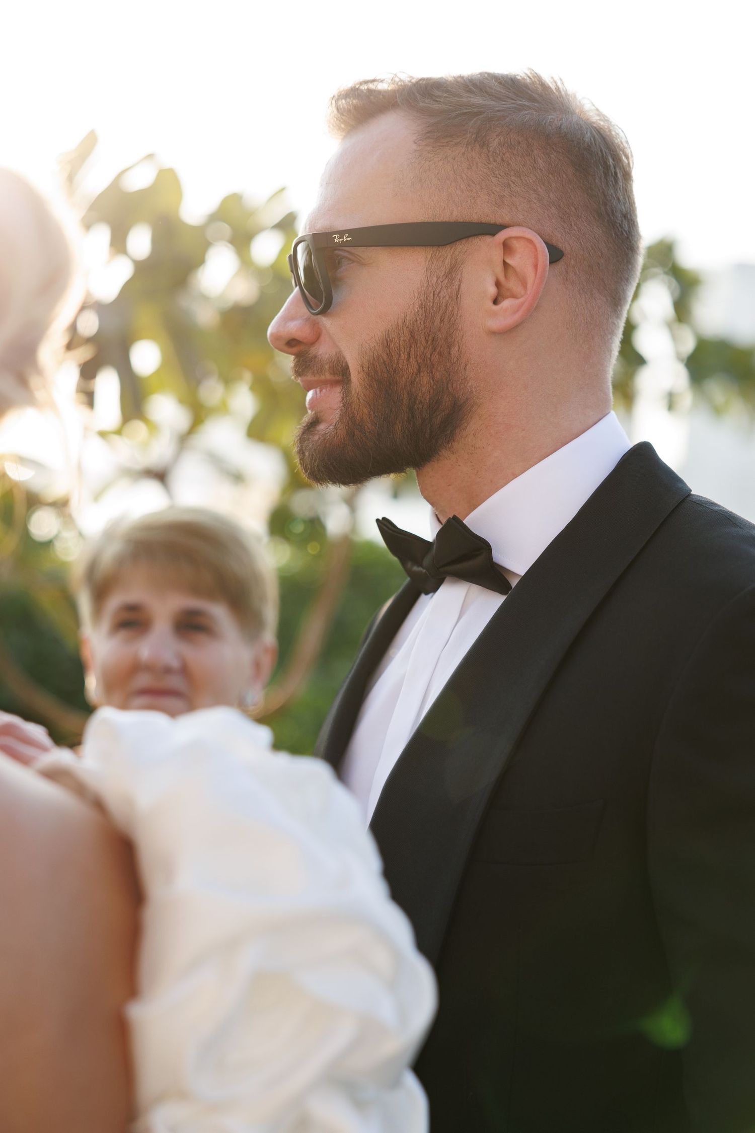 Formal wedding party members in black tuxedos and sunglasses gather outdoors.