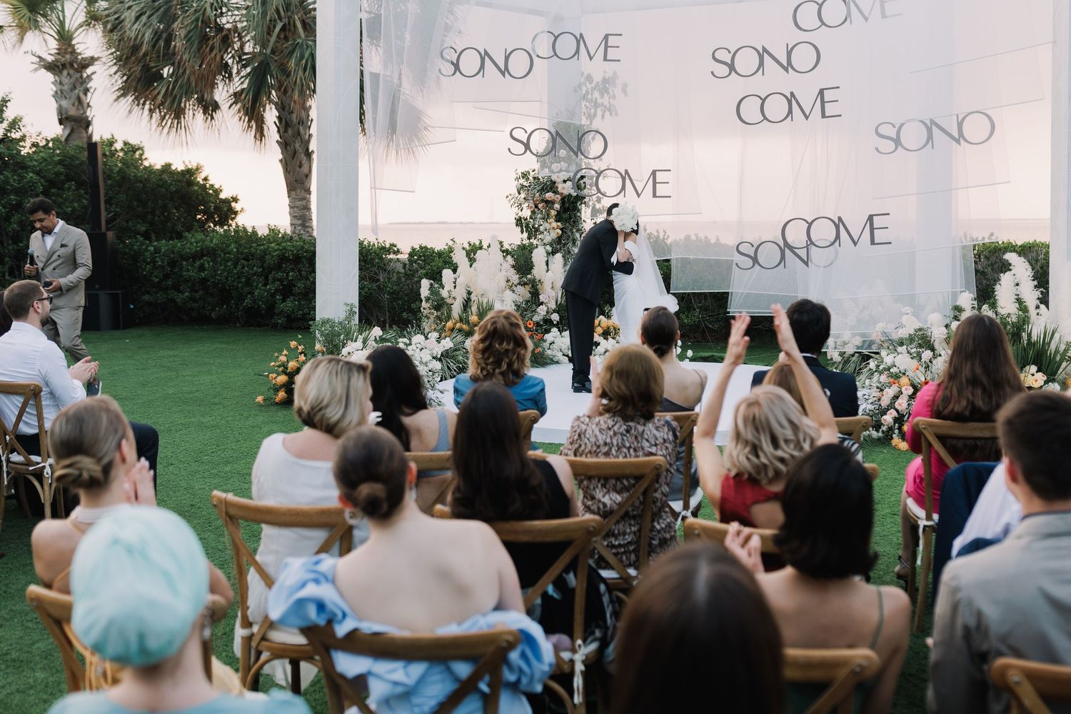 Wide angle view of wedding guests seated before a white backdrop with text.