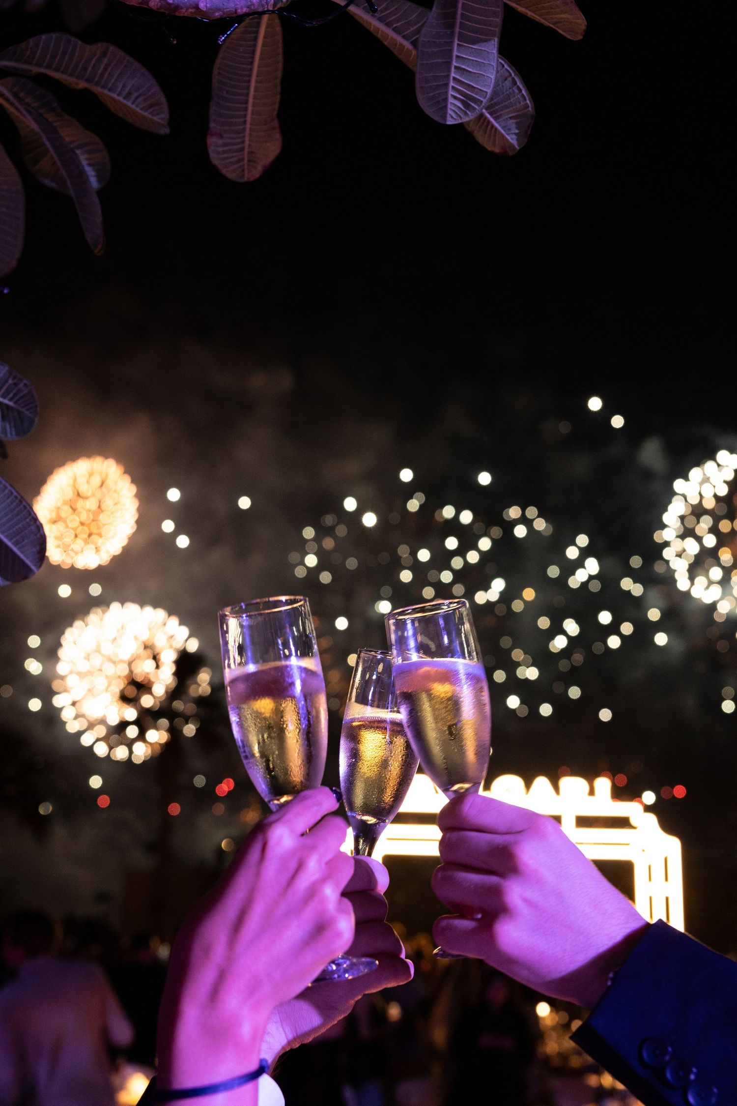 Hands raise champagne glasses against a backdrop of sparkling fireworks in a celebratory toast.