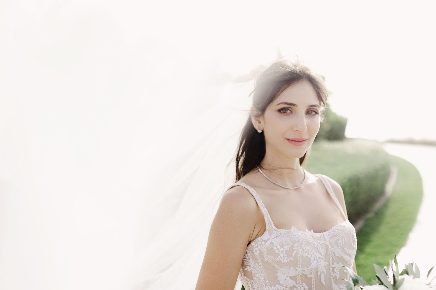 Elegant bride in a white lace dress poses outdoors in natural lighting.
