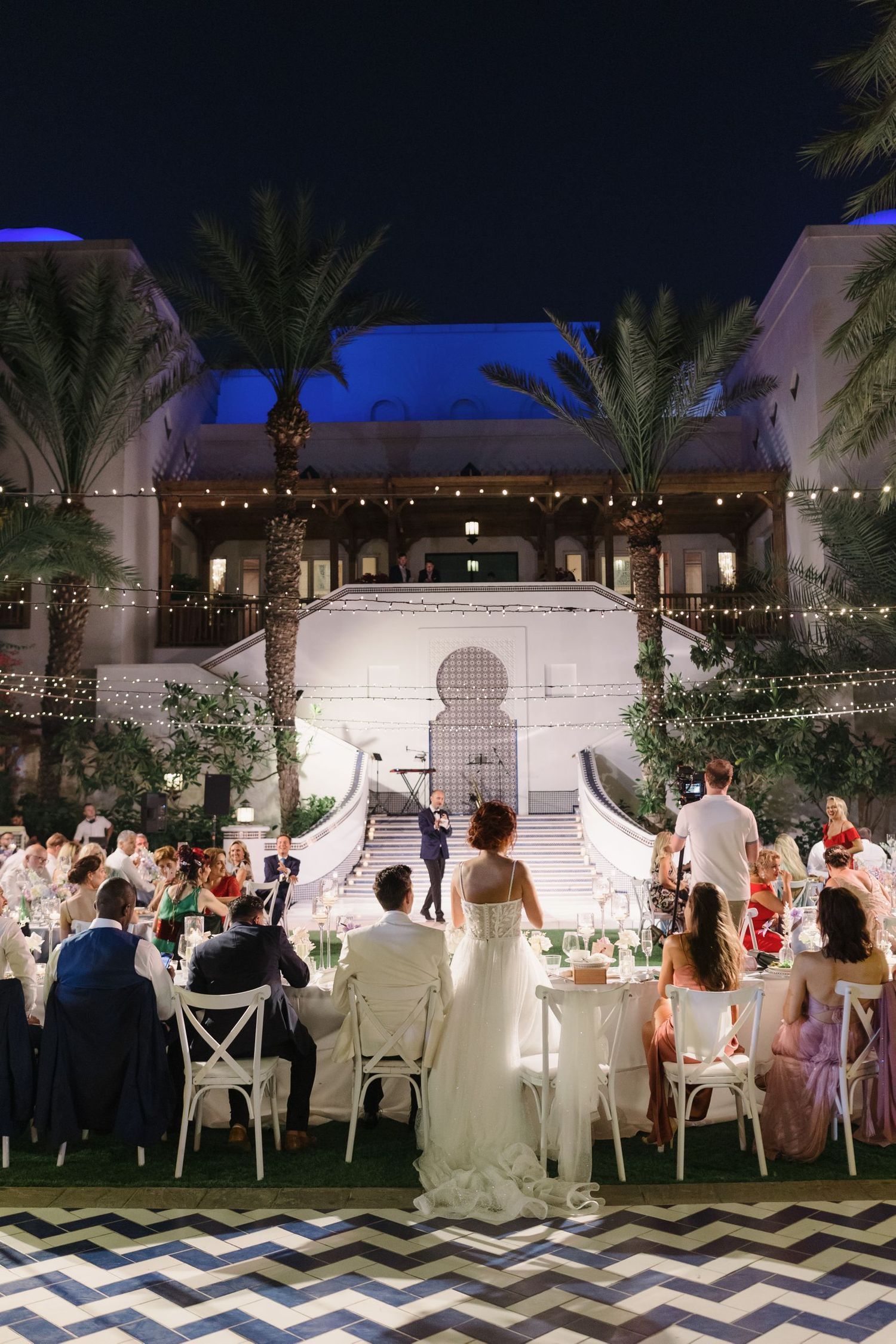 Panoramic view of an outdoor wedding reception with string lights and palm trees at dusk.