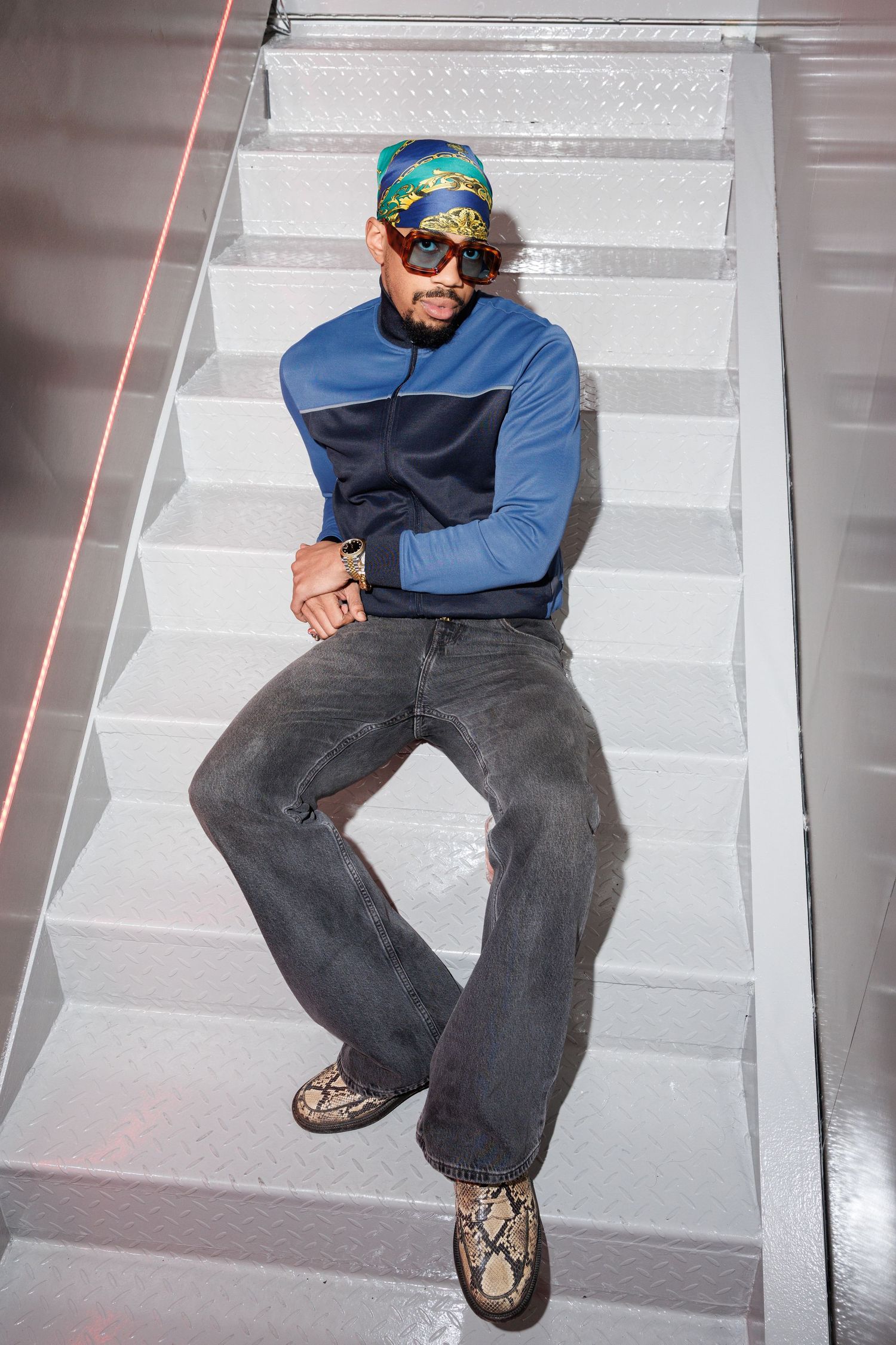 Person in blue jacket and sunglasses sits casually on white stairs wearing dark jeans and sneakers.