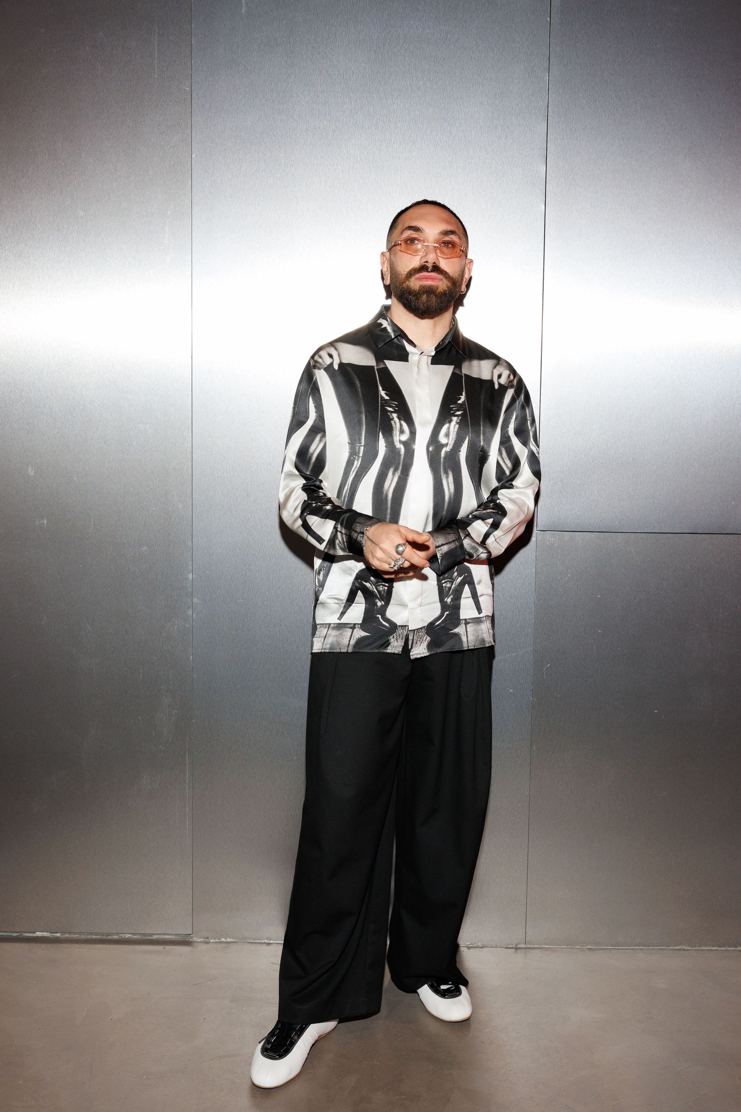 Individual in striped jacket and black pants poses casually against grey studio wall.