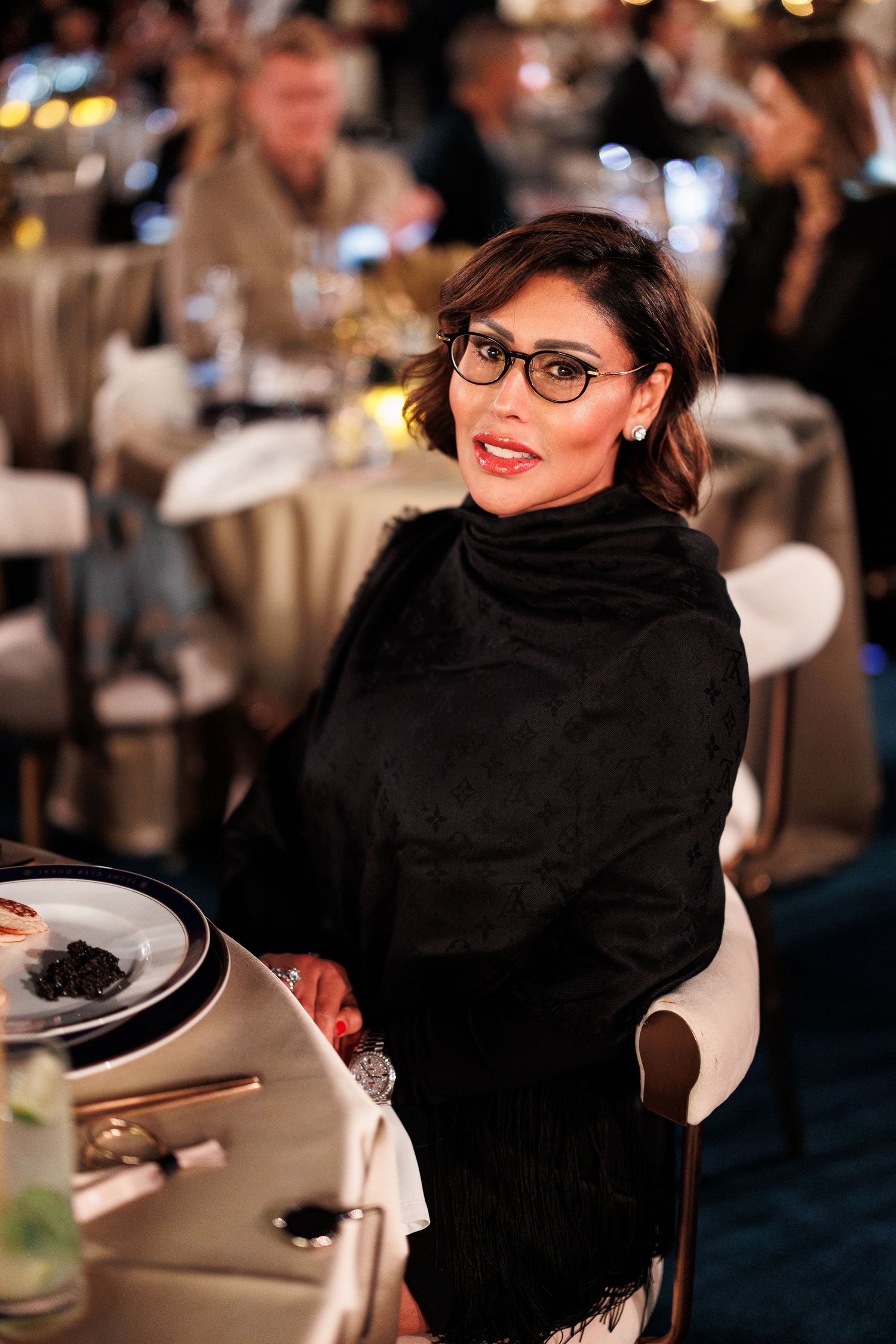 A person in black attire and glasses smiles while sitting at a formal dining event with blurred background lighting.
