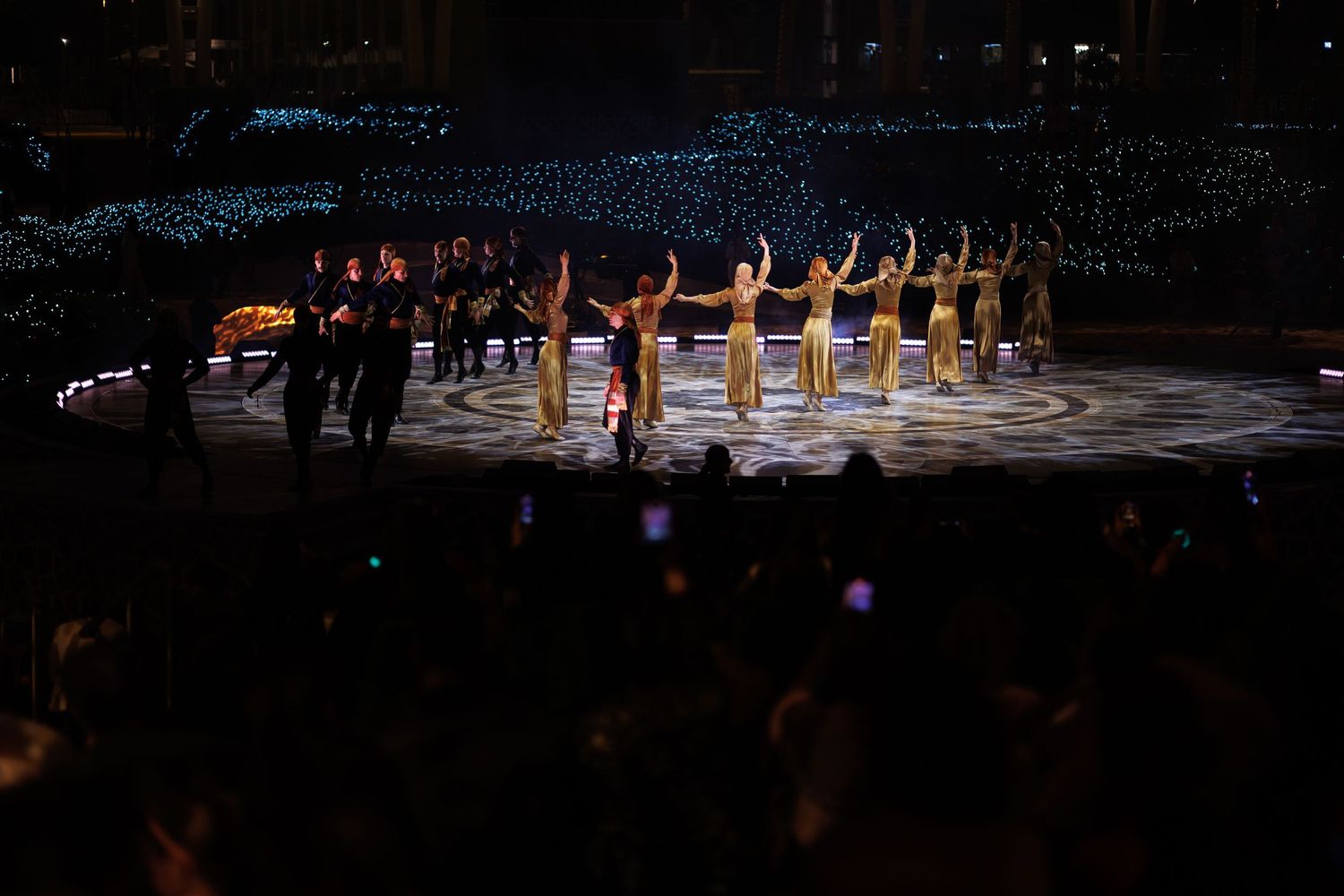 Performers lined up on stage with blue lighting effects and audience silhouettes in foreground.
