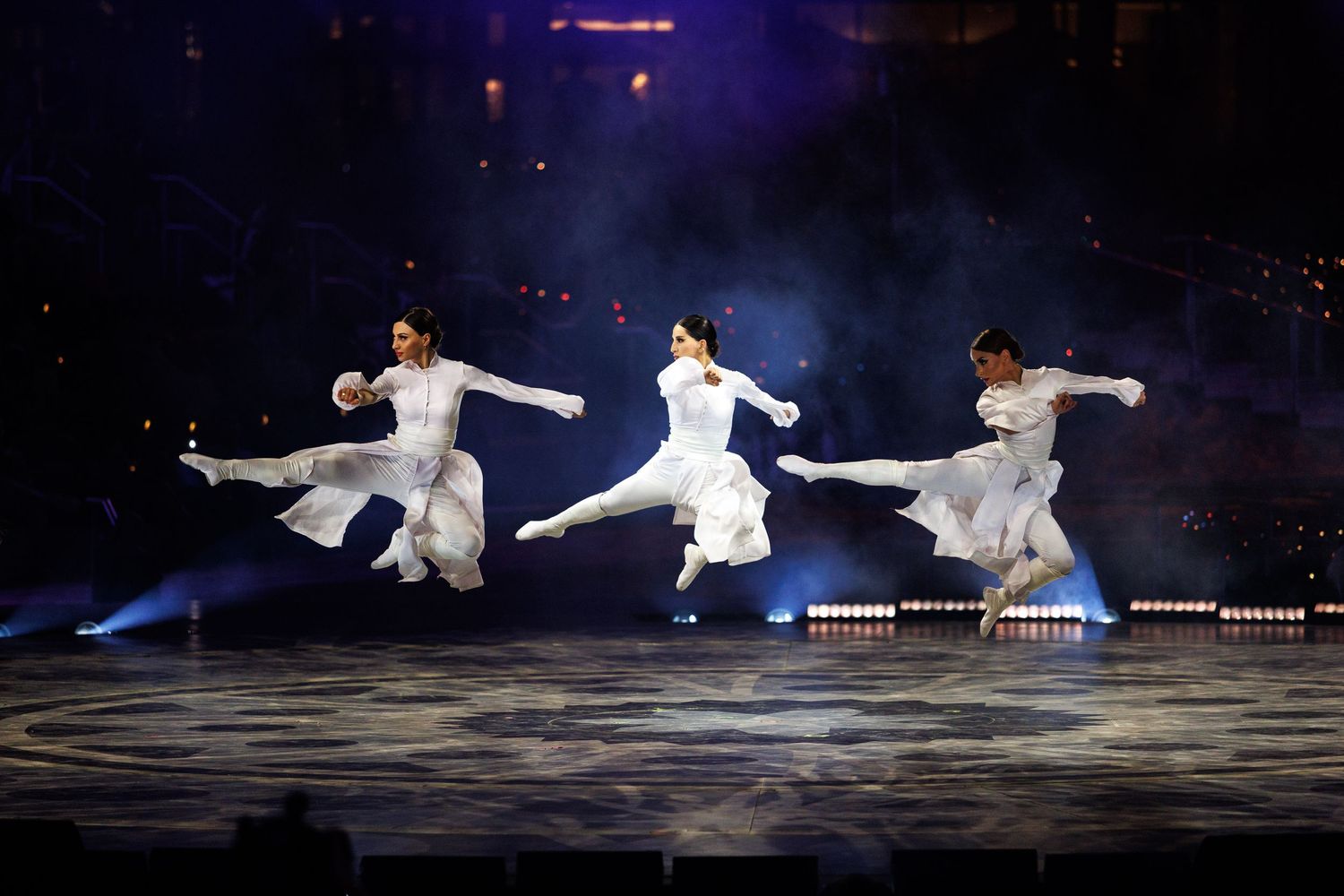 Figure skaters perform graceful jumps and spins on ice during an evening performance with blue lighting.