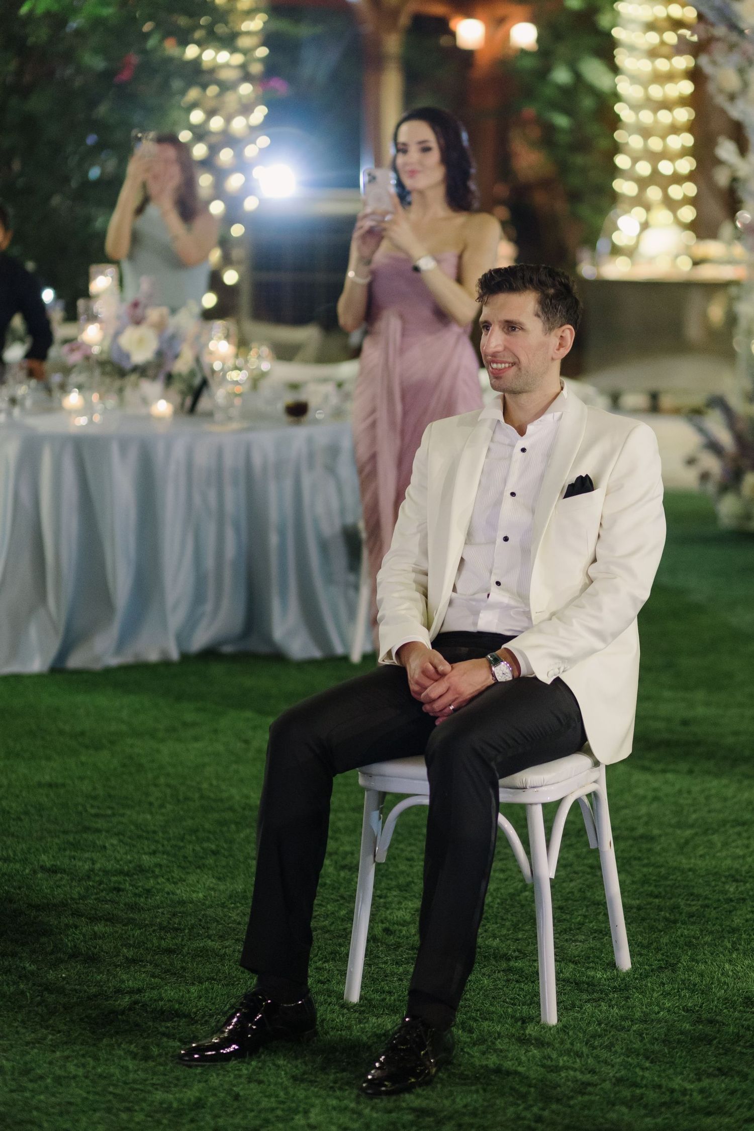 Guest in white jacket sits on chair during outdoor evening wedding reception.