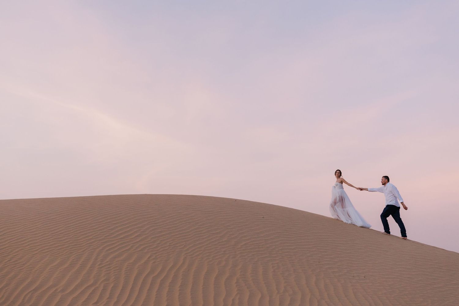 Silhouettes of people walking along sand dunes against pastel sky.