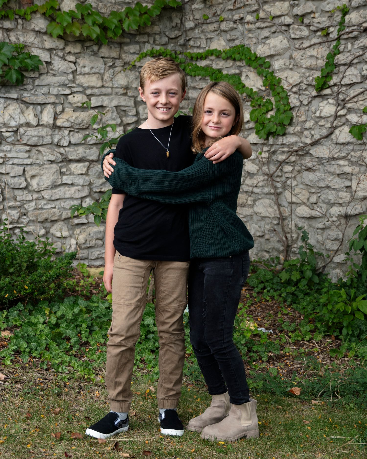 Two young people pose together against a stone wall covered with ivy.