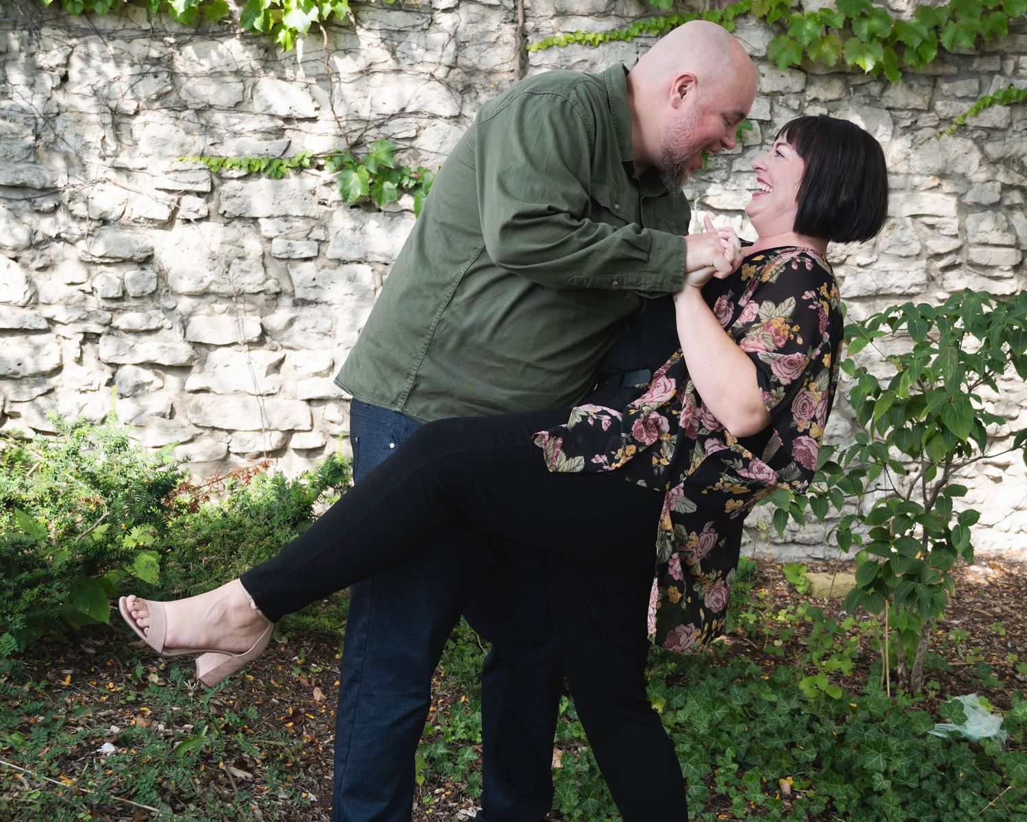 A couple shares a tender moment against a stone wall in an outdoor garden setting.