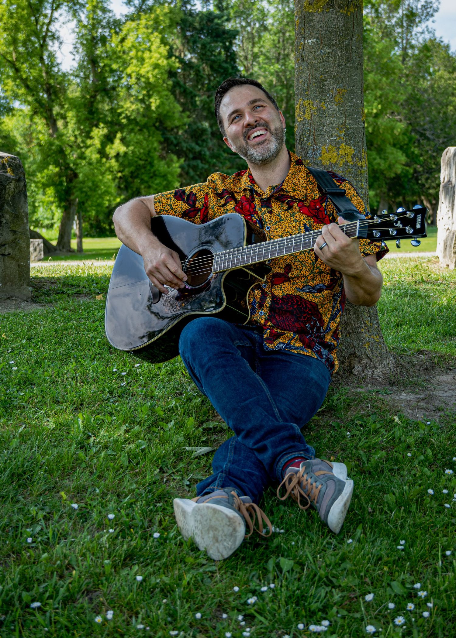 Person playing acoustic guitar while sitting casually under trees in park setting.