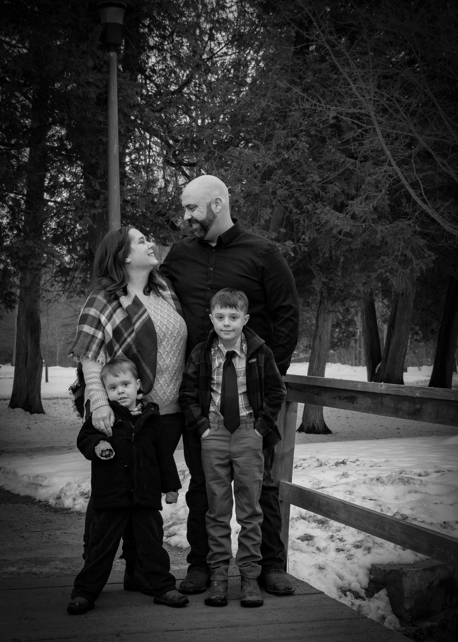 Family photo session in a snowy winter park with black and white filter showing candid moments on a wooden walkway.