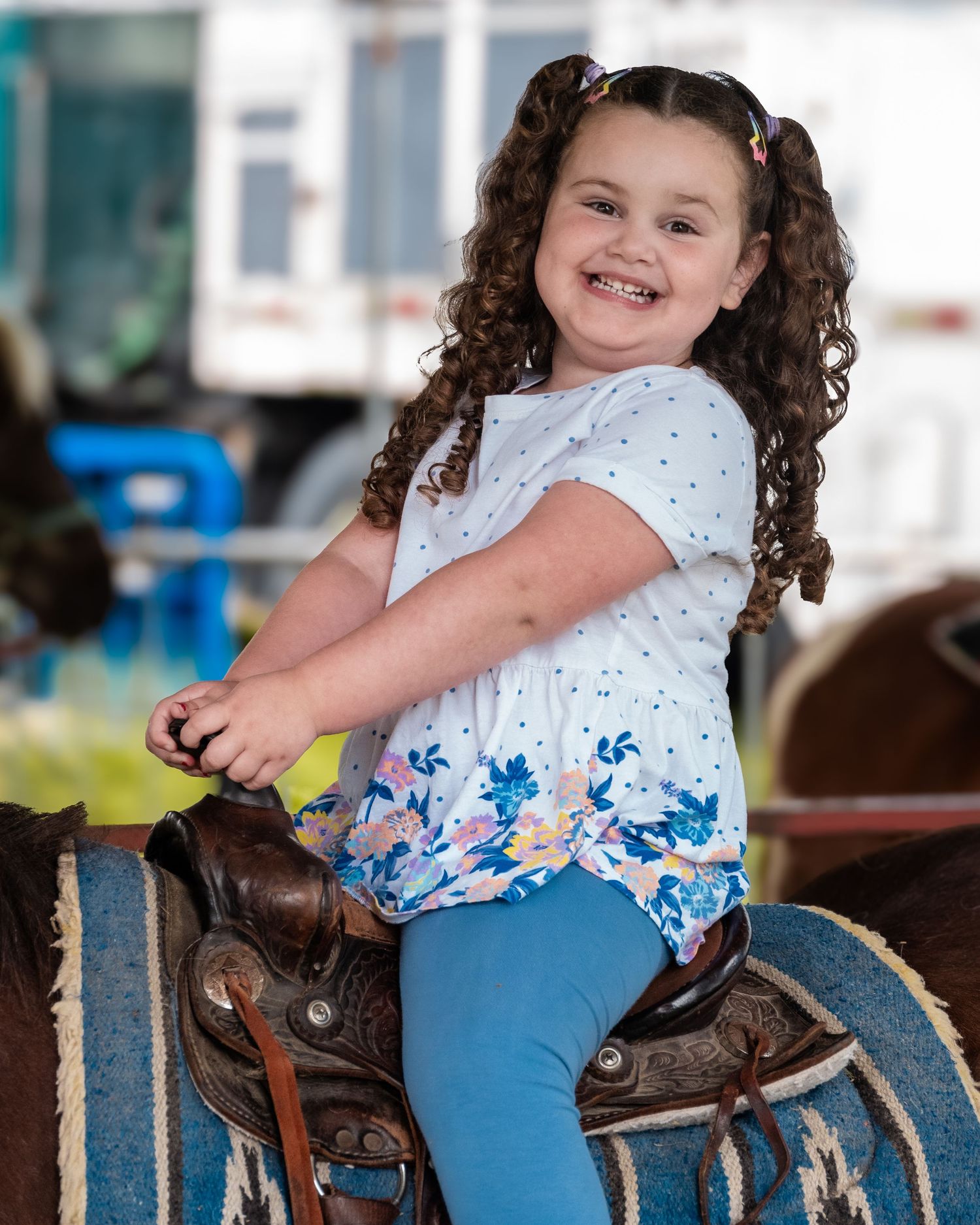 Young child in blue and white dress sitting on western saddle at horse stables.