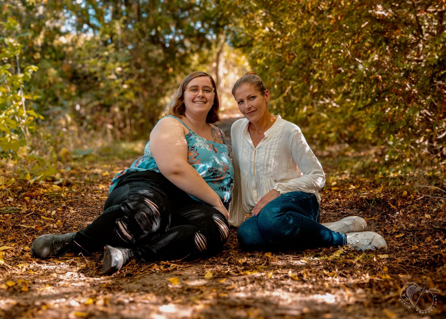 Two people share laughter while sitting on a leaf-covered forest path.
