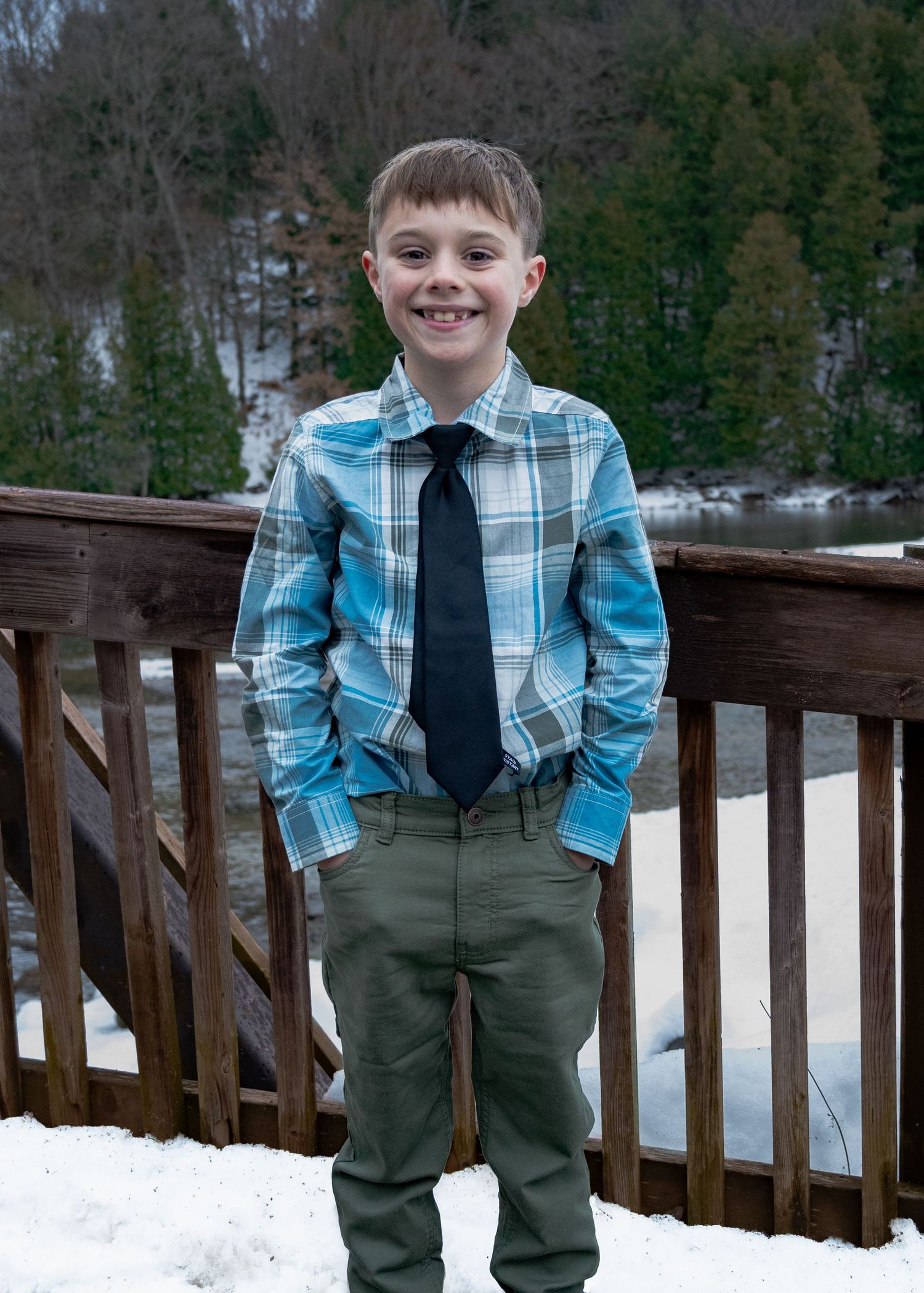 Child wearing plaid shirt and tie poses on wooden railing during winter photoshoot.