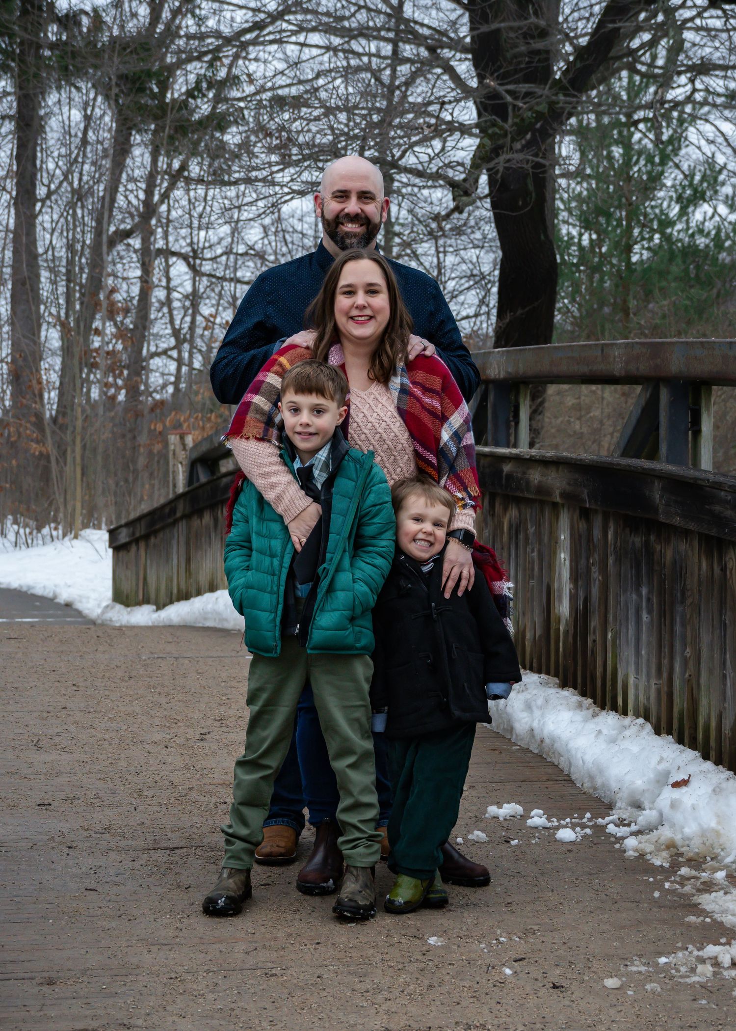 Family enjoying a winter photoshoot on a wooden bridge surrounded by snow and trees.