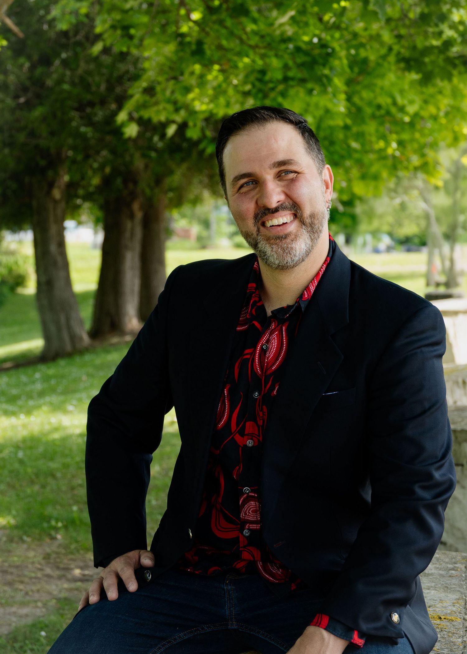 Person in black blazer and red patterned shirt poses against green foliage in outdoor setting.