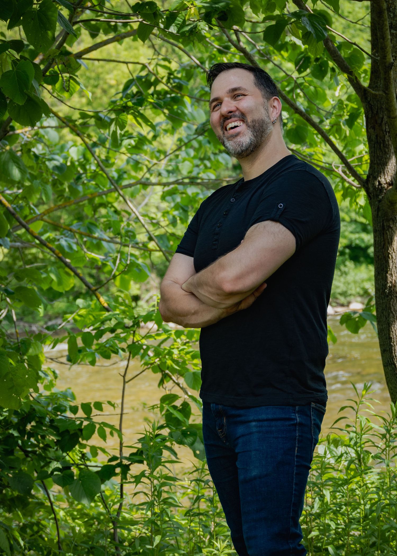 Person in black shirt poses confidently with crossed arms against leafy background.