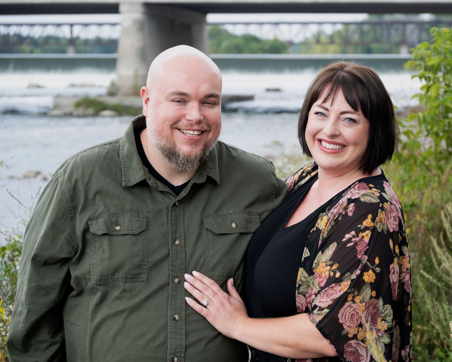 A happy couple poses together outdoors wearing casual attire near a waterfront.