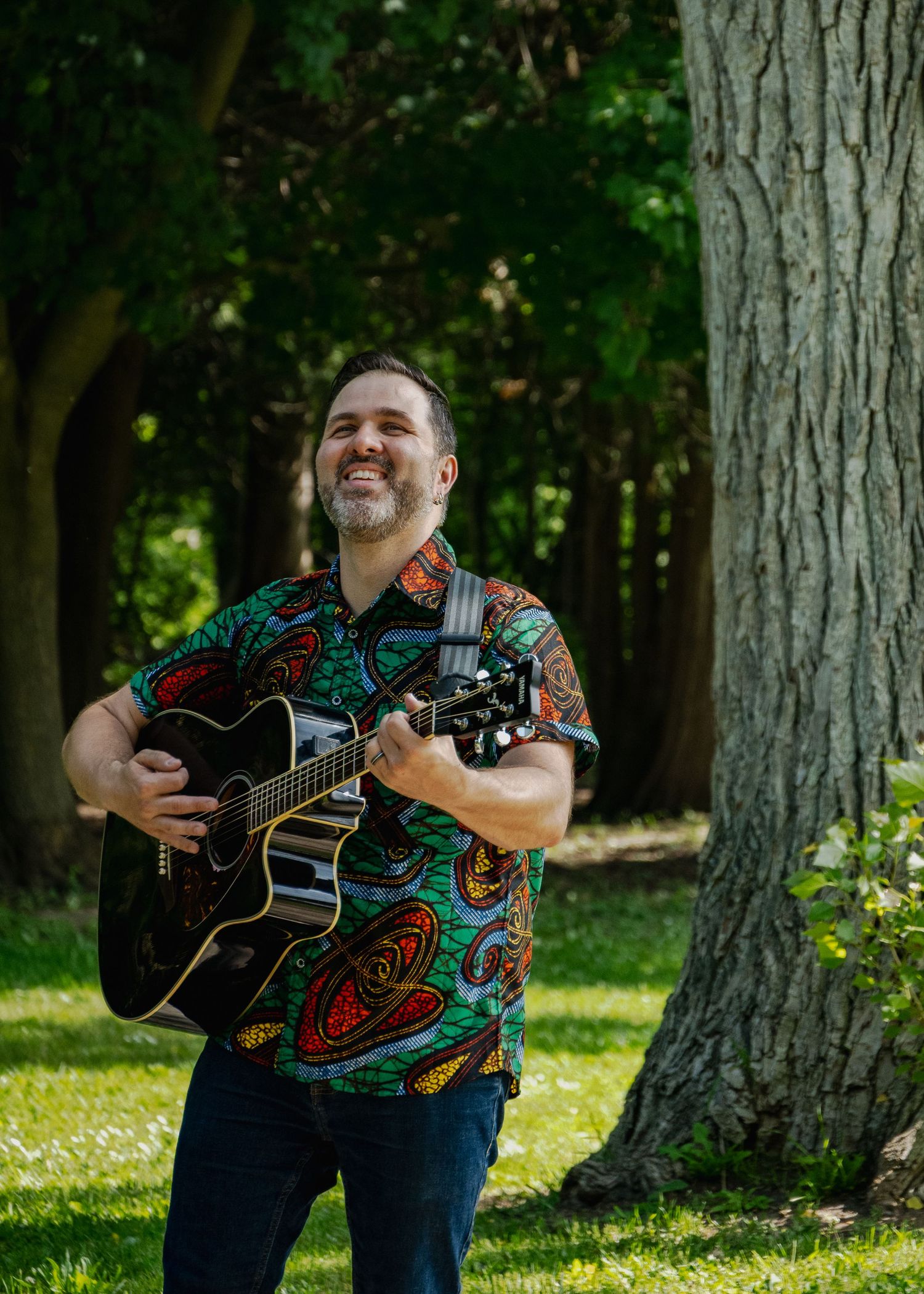 Musician playing acoustic guitar in a wooded setting with dappled sunlight.