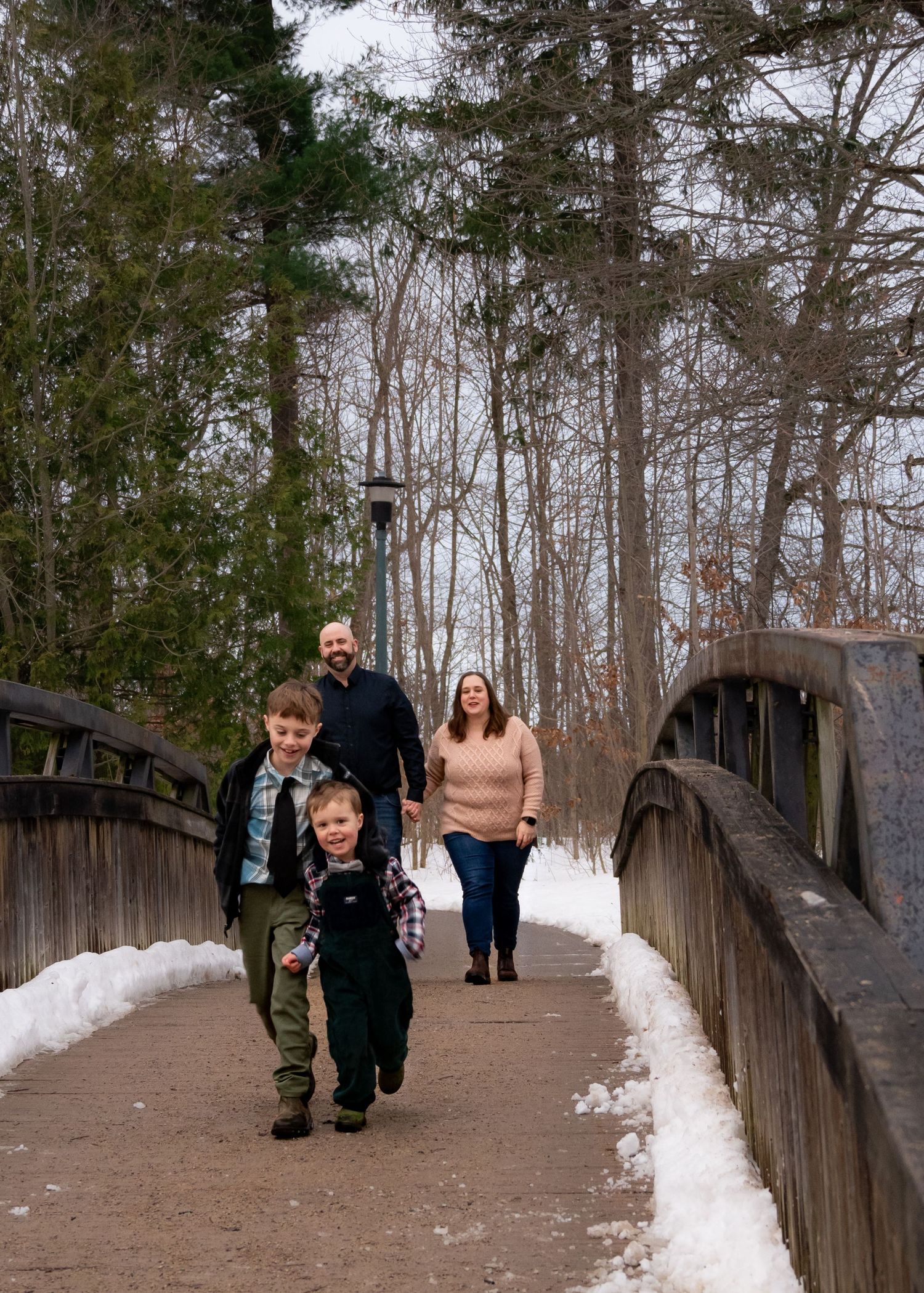 People walking along a snowy wooden bridge pathway through trees in winter.
