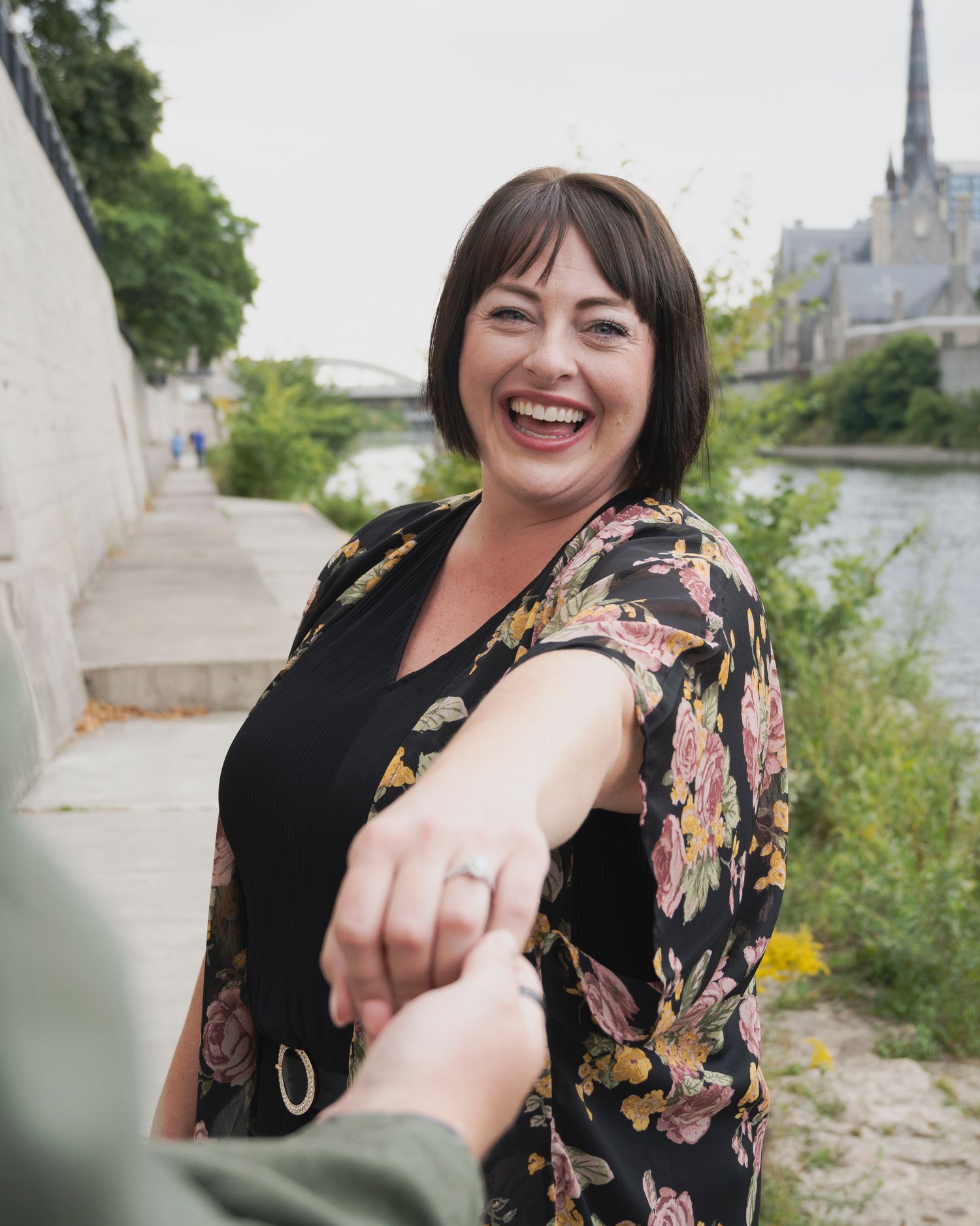 Happy couple celebrating their engagement by a riverside with a church spire in the background.