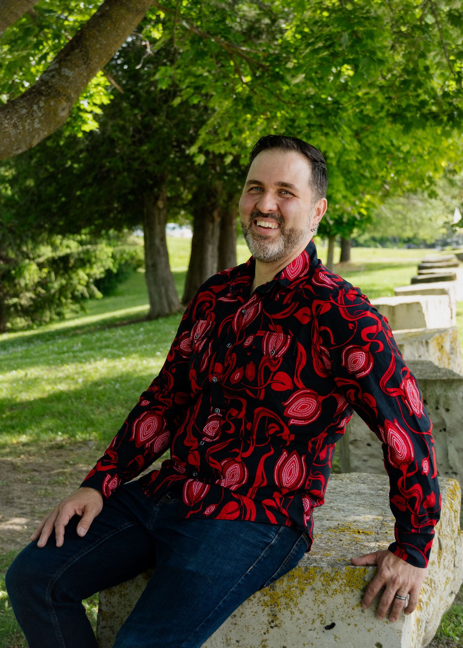 Person wearing black and red floral shirt sits casually on stone bench in park setting.