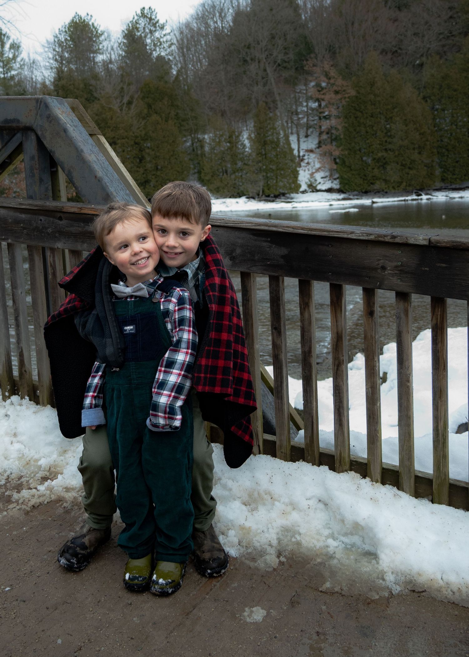 Two children posing together on a snowy winter walkway surrounded by trees.