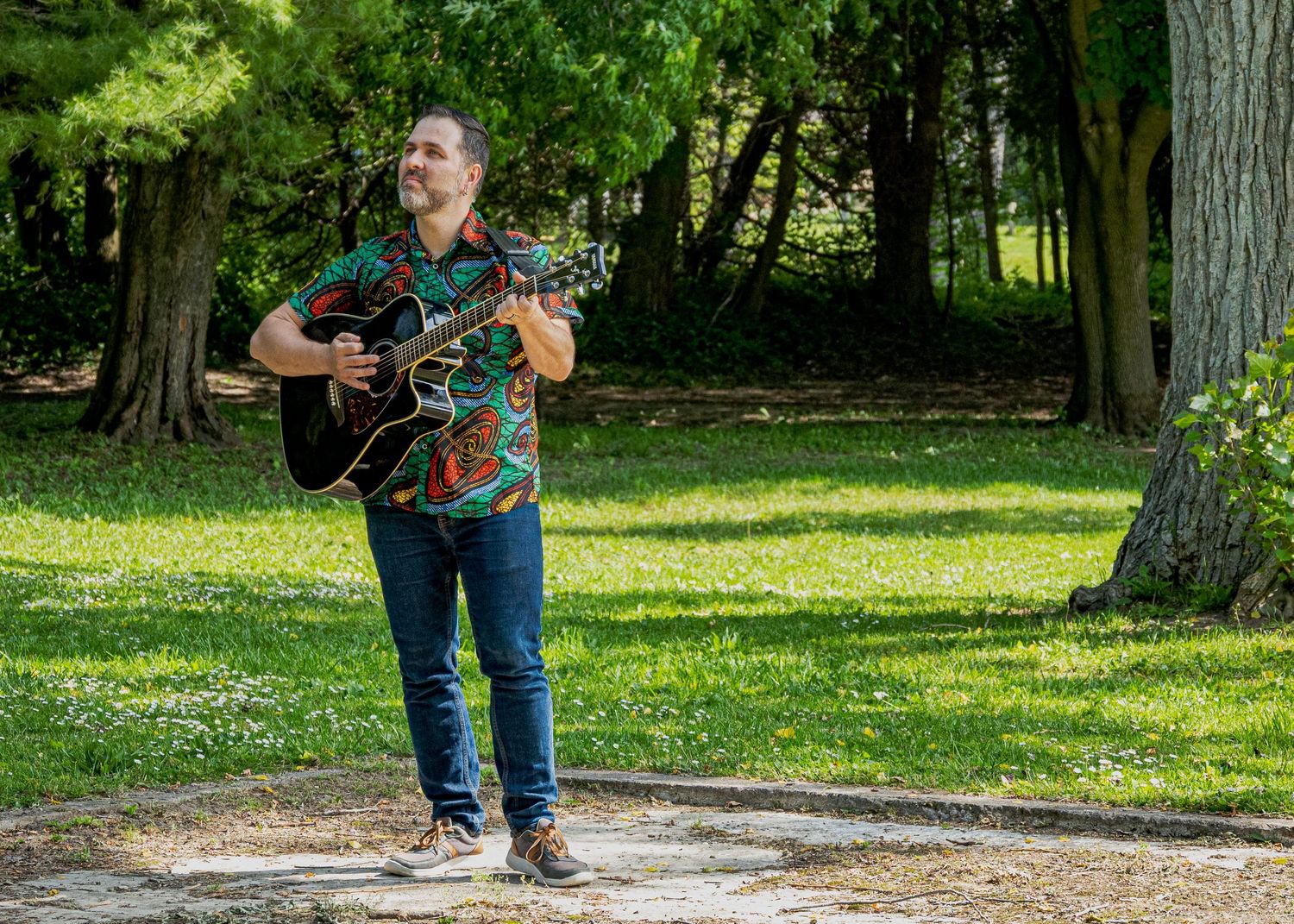 Person with acoustic guitar posing in a sunny park setting with tall trees.