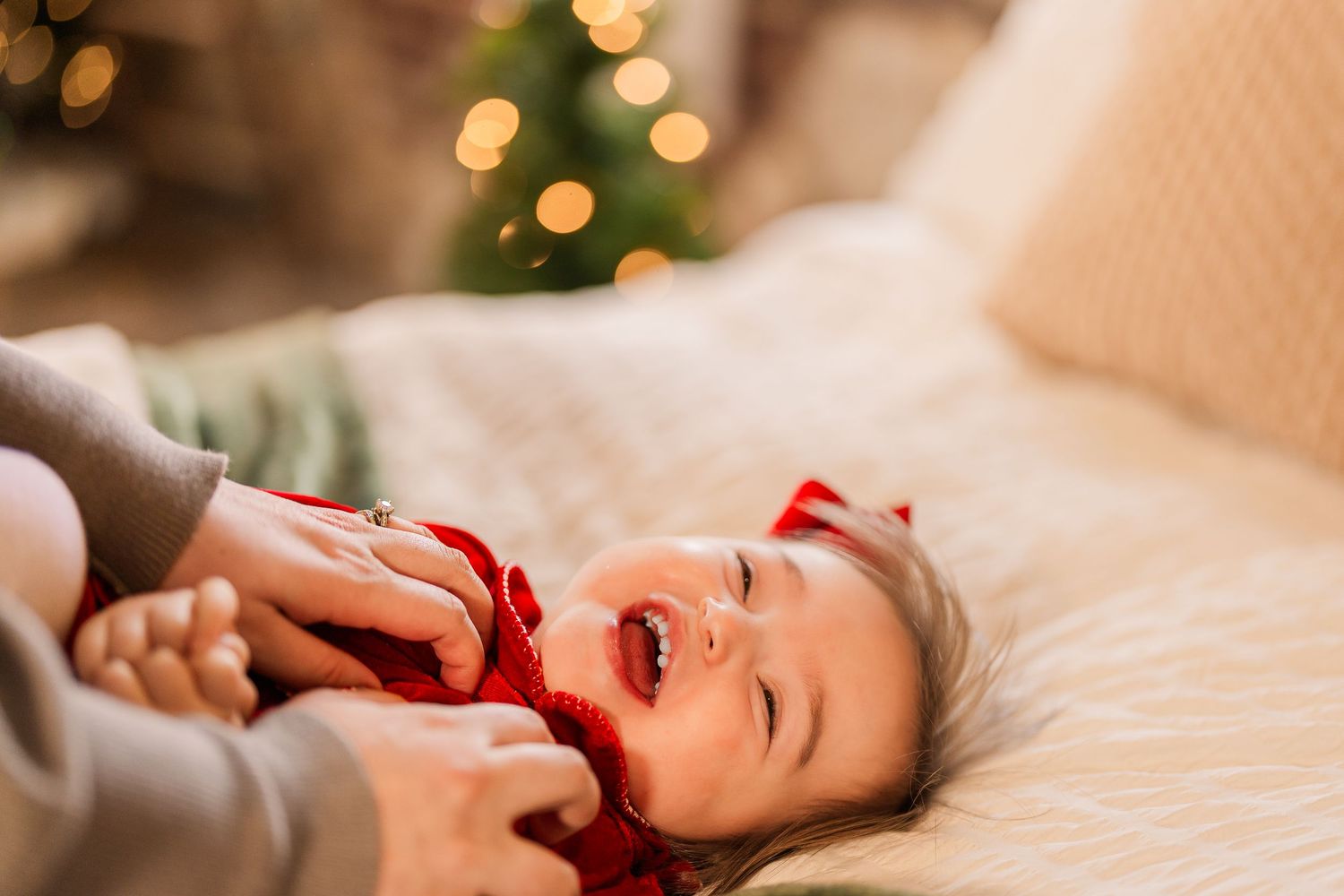 Greensboro, NC Family Photographer, Ryan Harden, Reidsville NC, toddler girl laughs on bed as mom tickles with Christmas tree in background.