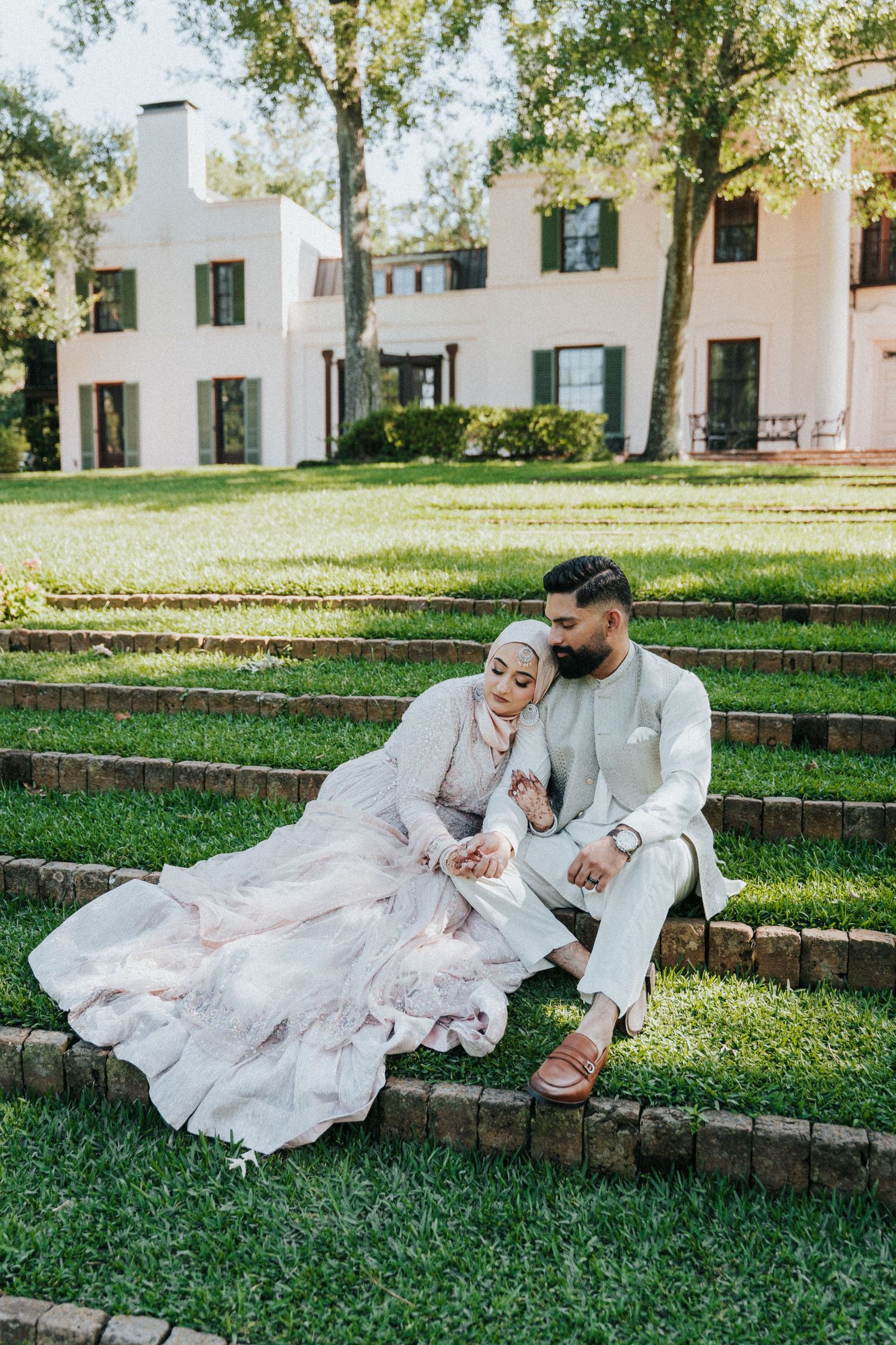 Muslim bride and groom in elegant wedding attire sitting on garden steps before a white manor house.