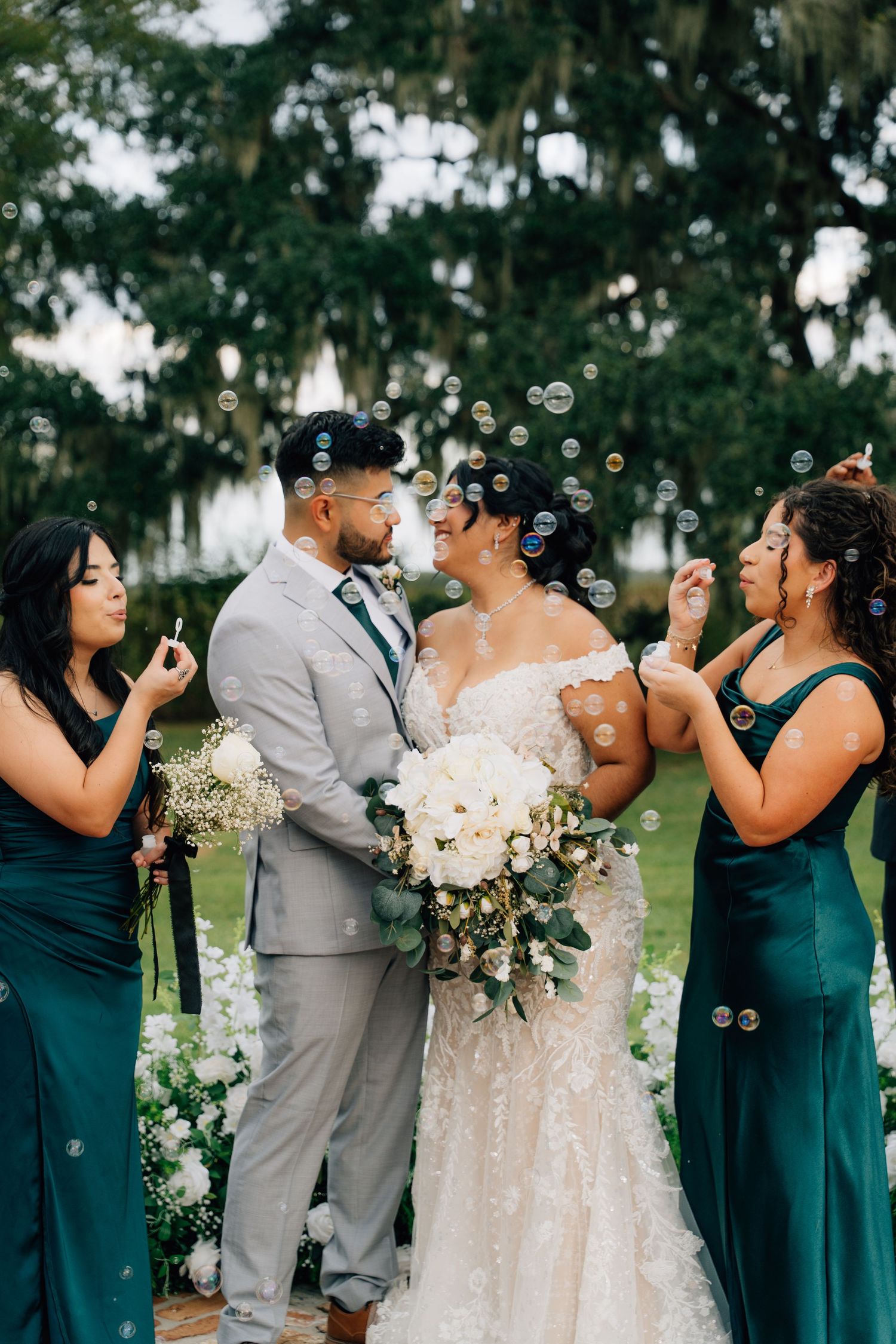 Wedding party in teal dresses and gray suits blowing bubbles together in a joyful outdoor celebration.