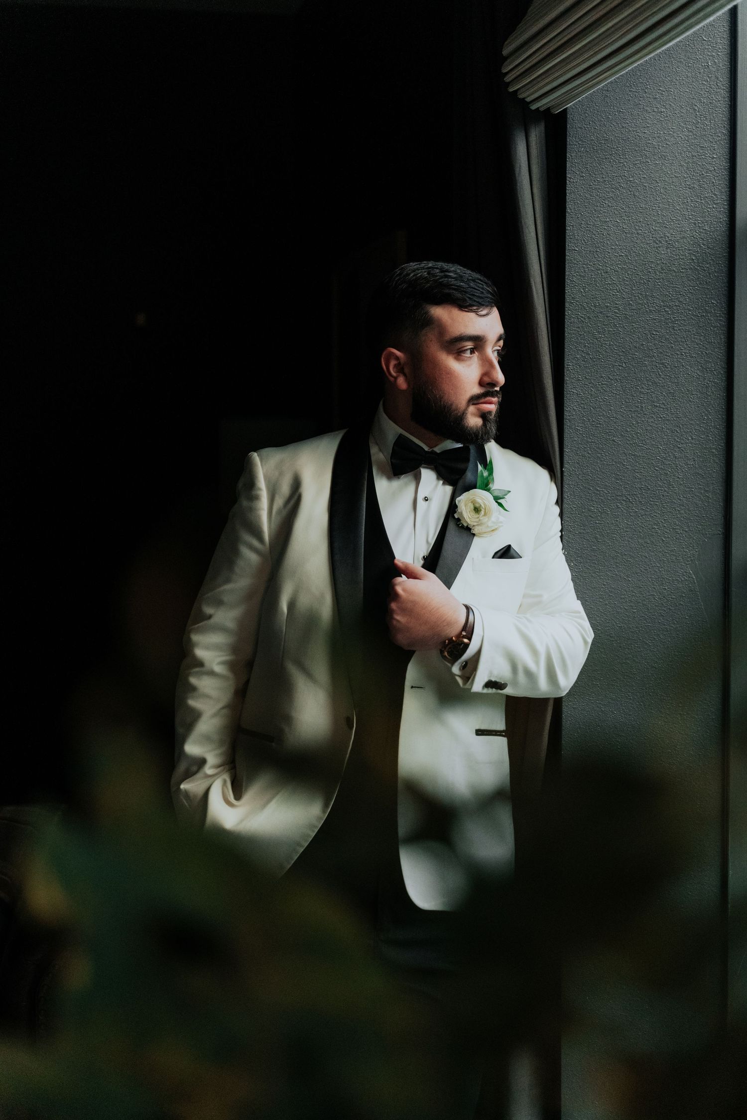 Groom in white tuxedo jacket adjusts his bowtie while standing in dramatic lighting against dark background.