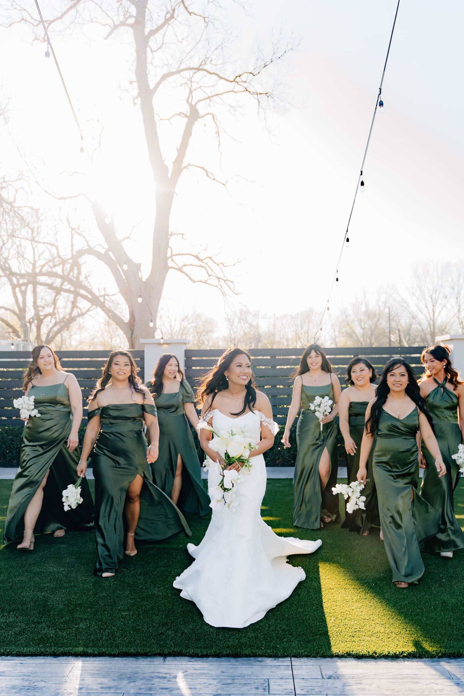 Bridesmaids in emerald green dresses pose playfully on grass with white bouquets.