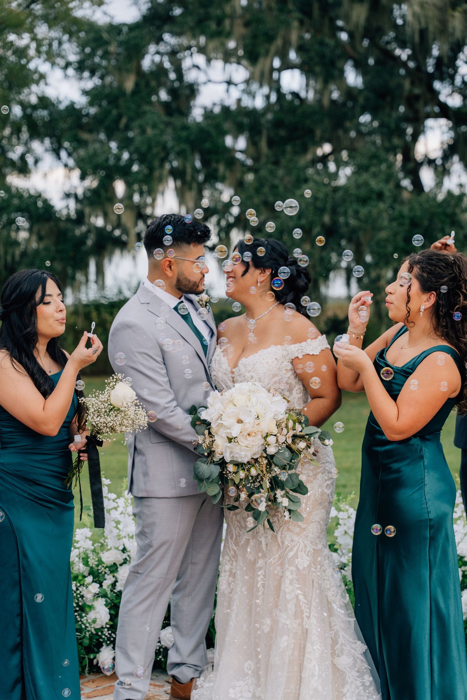Wedding party blowing bubbles during outdoor ceremony with bridesmaids in teal dresses.