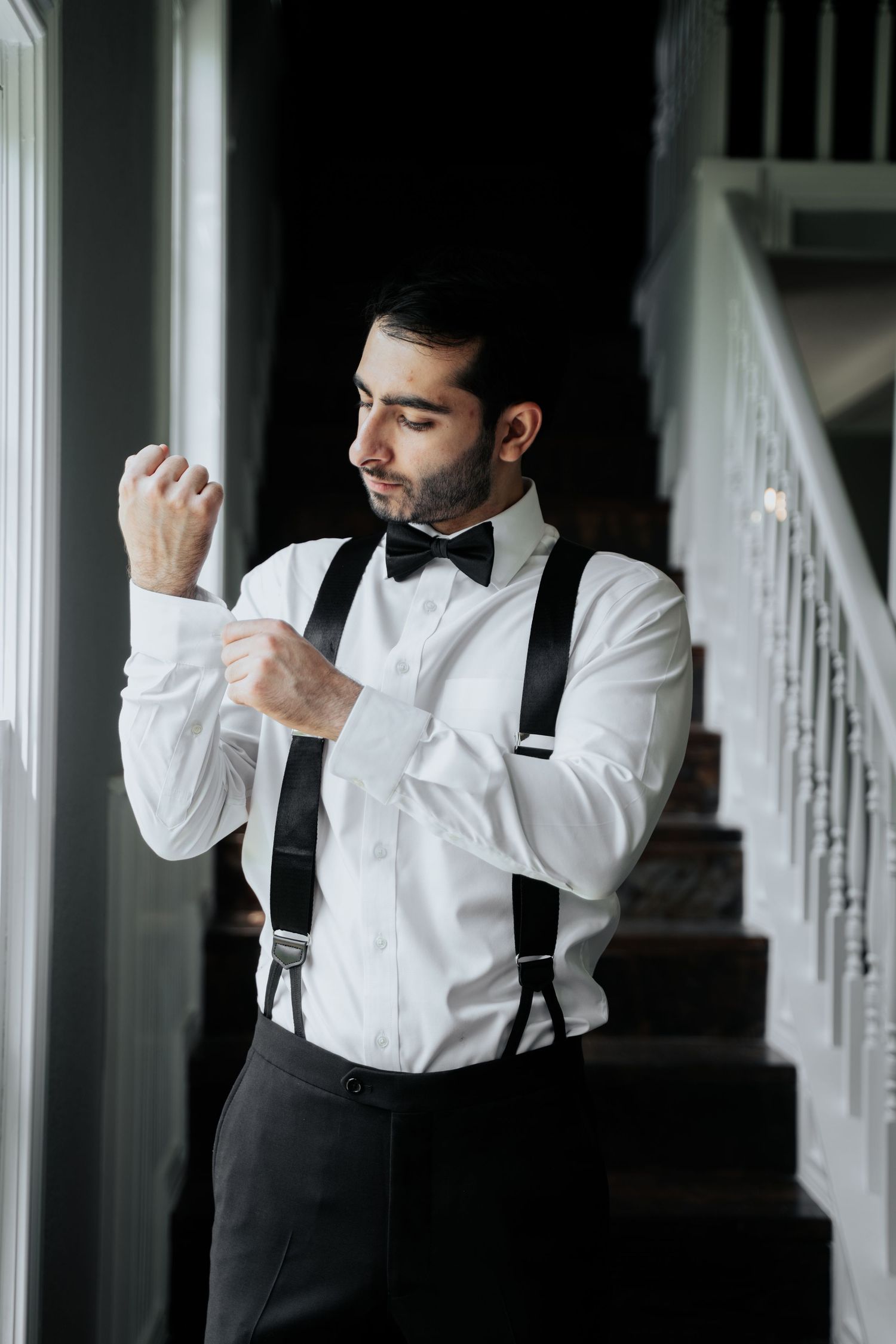 Artistic series of groom getting ready in white shirt and suspenders against white staircase.