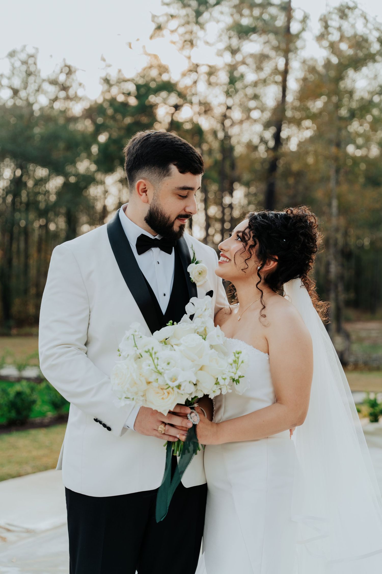 A romantic wedding couple shares an intimate moment while holding a white bouquet in an outdoor setting.