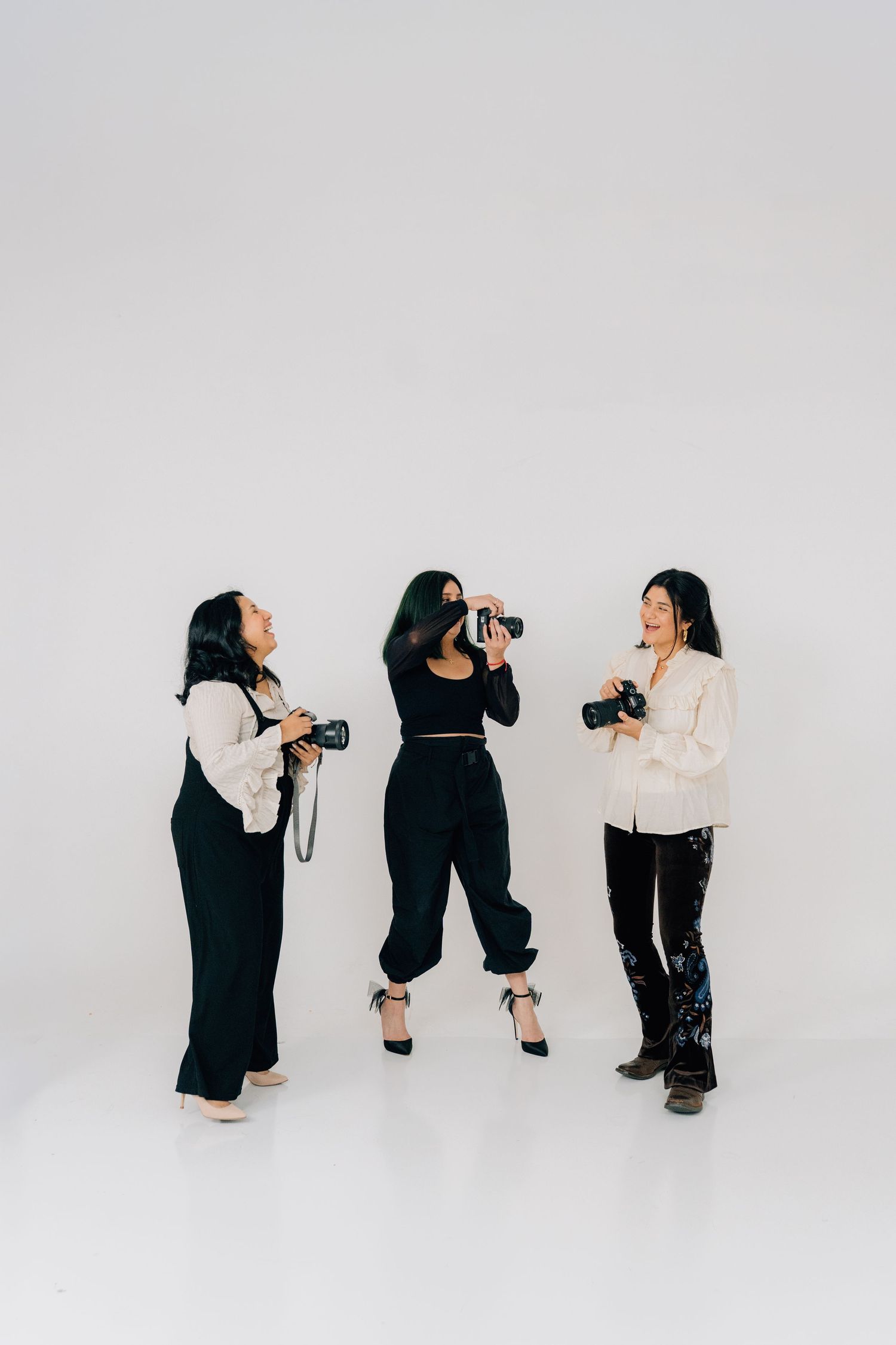 Three photographers in dark clothing take pictures of each other in a white studio setting.