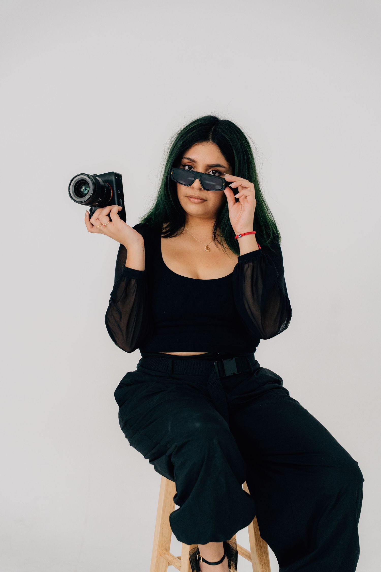 Photographer in black outfit poses with camera and sunglasses on wooden stool.