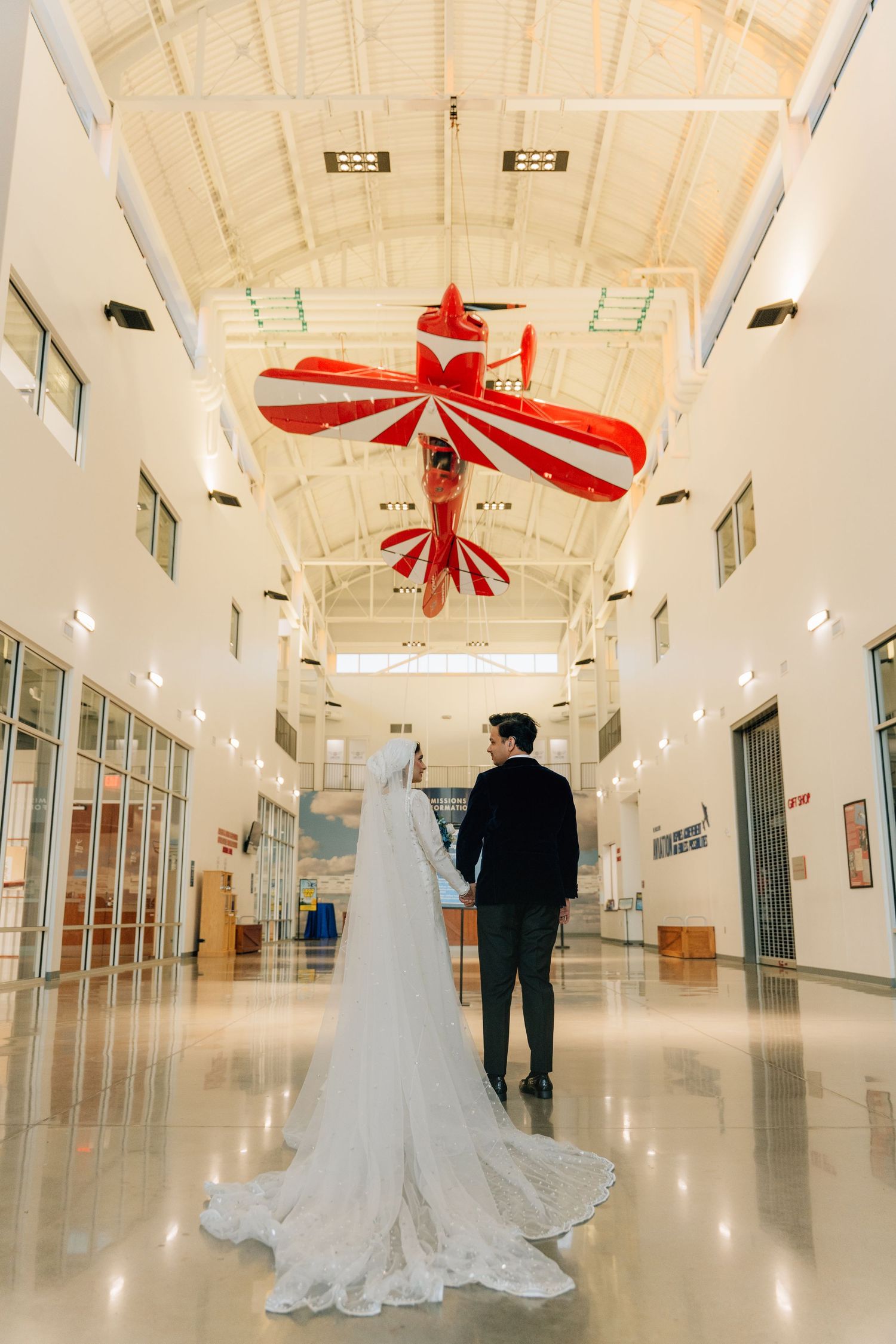Wedding couple walks down long white hallway with red vintage airplanes hanging from ceiling.