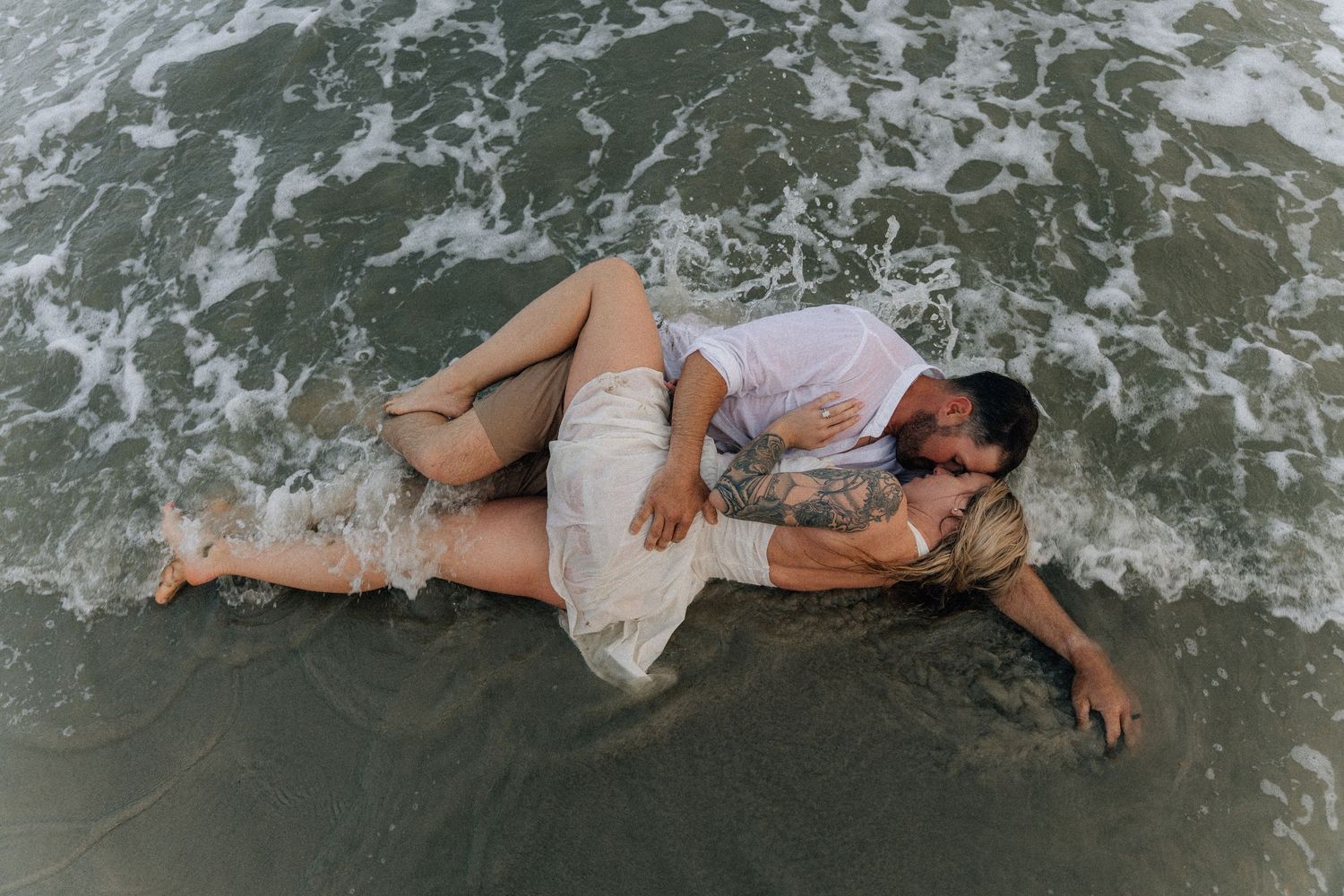 Couple lying in ocean waves during a beach photoshoot.