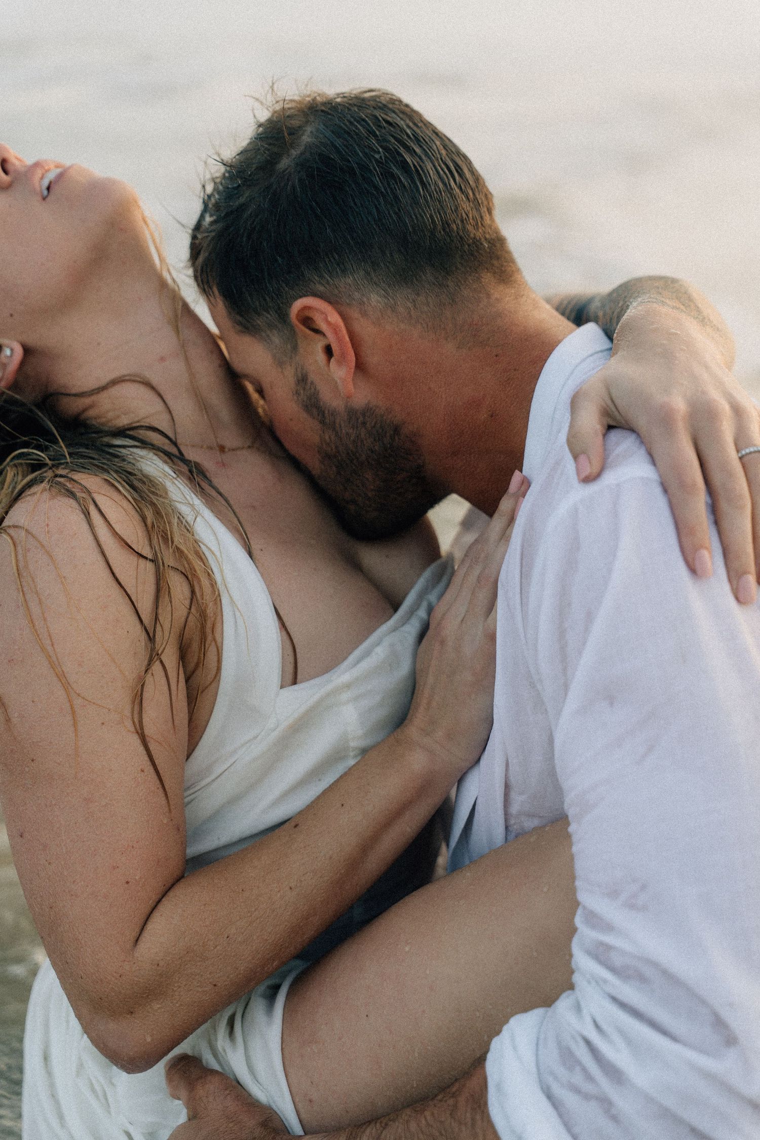 An intimate moment between a couple in white clothing embracing on a beach at sunset.
