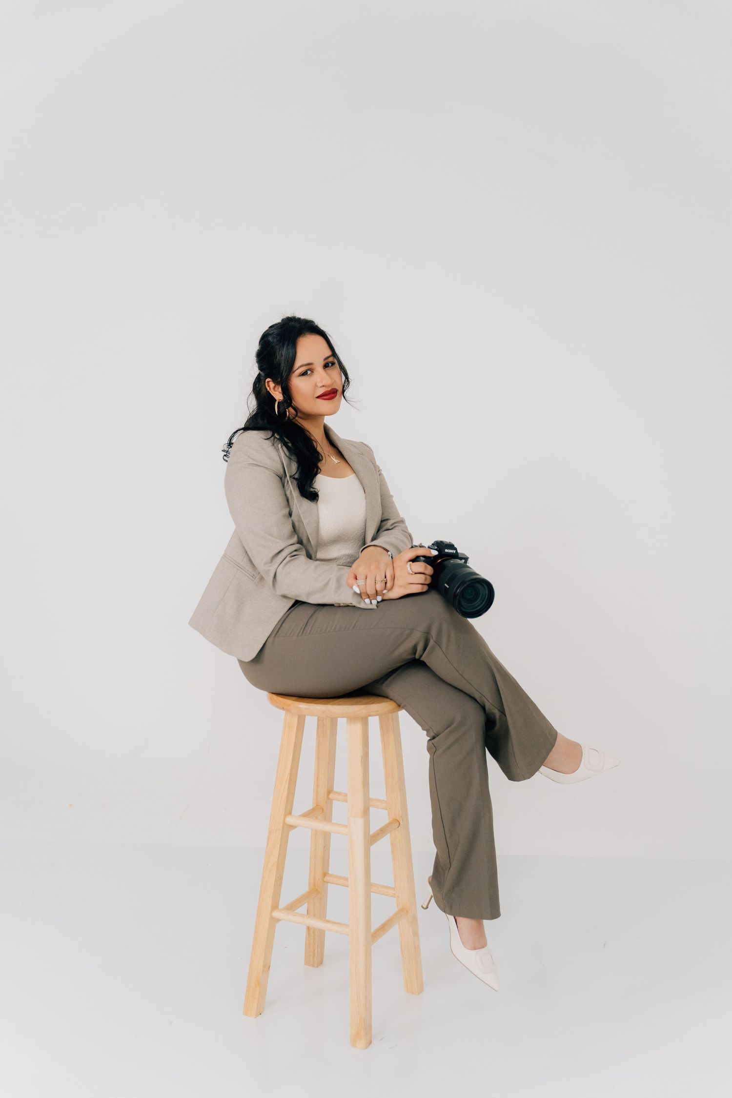 A professional woman in a beige suit sits on a wooden stool holding a camera against a white background.