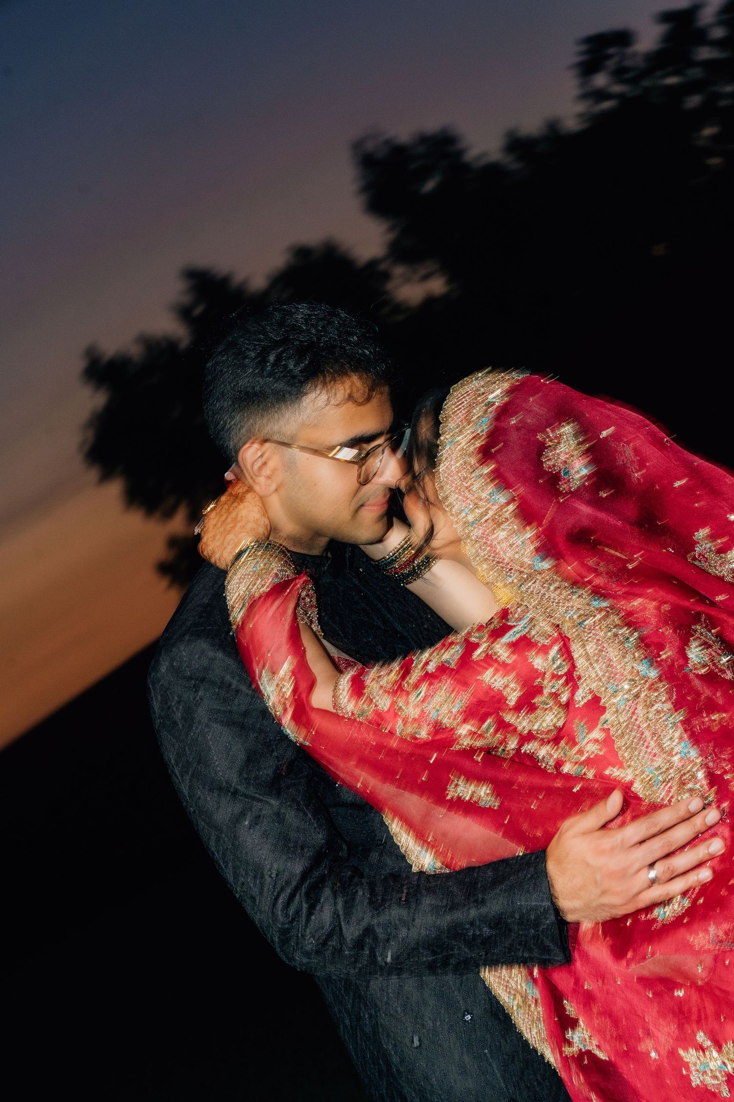 Couple in traditional Indian wedding attire sharing a romantic moment at dusk, bride in red lehenga.