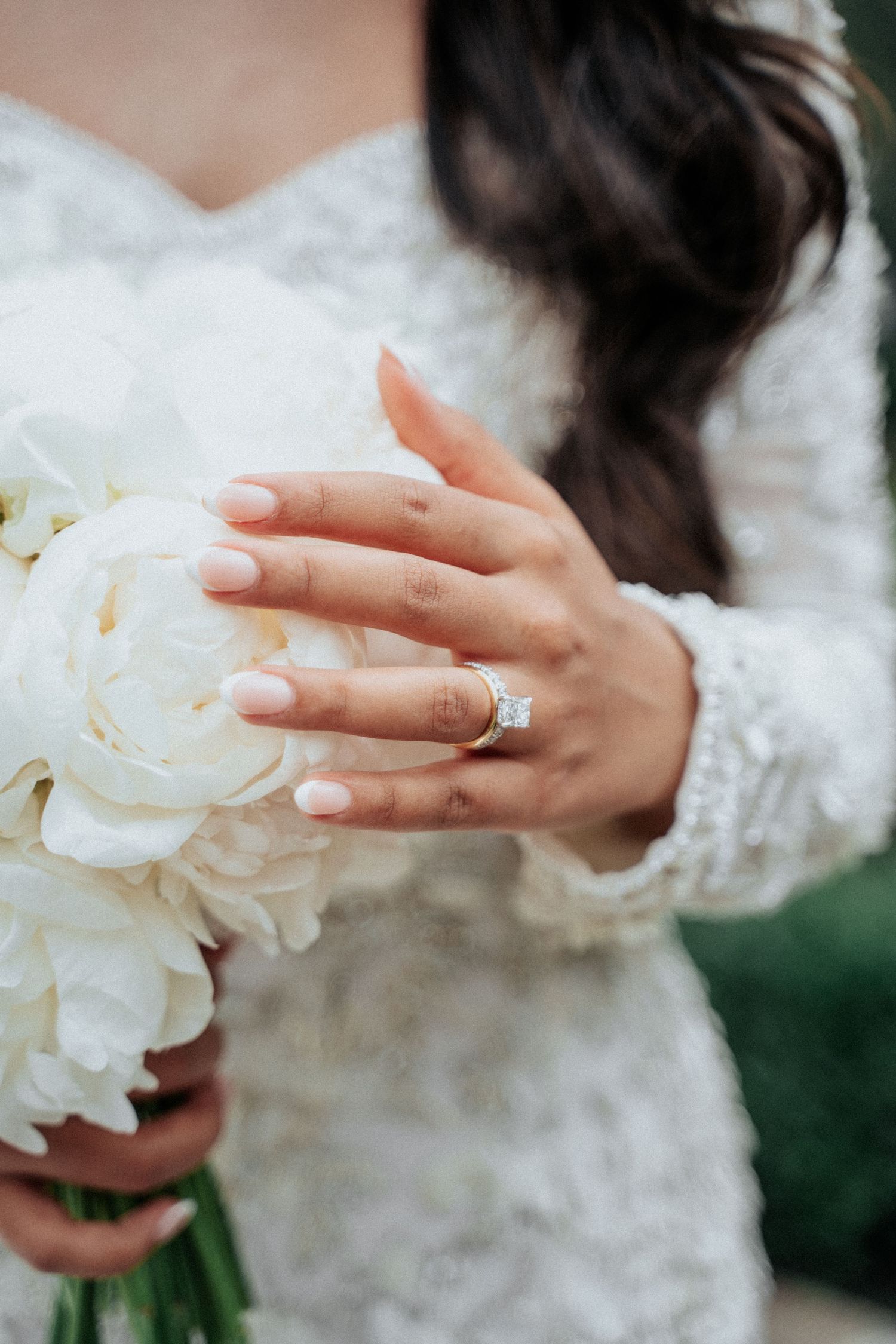 Close-up of a bride's hand with engagement ring holding white peonies against her lace dress.